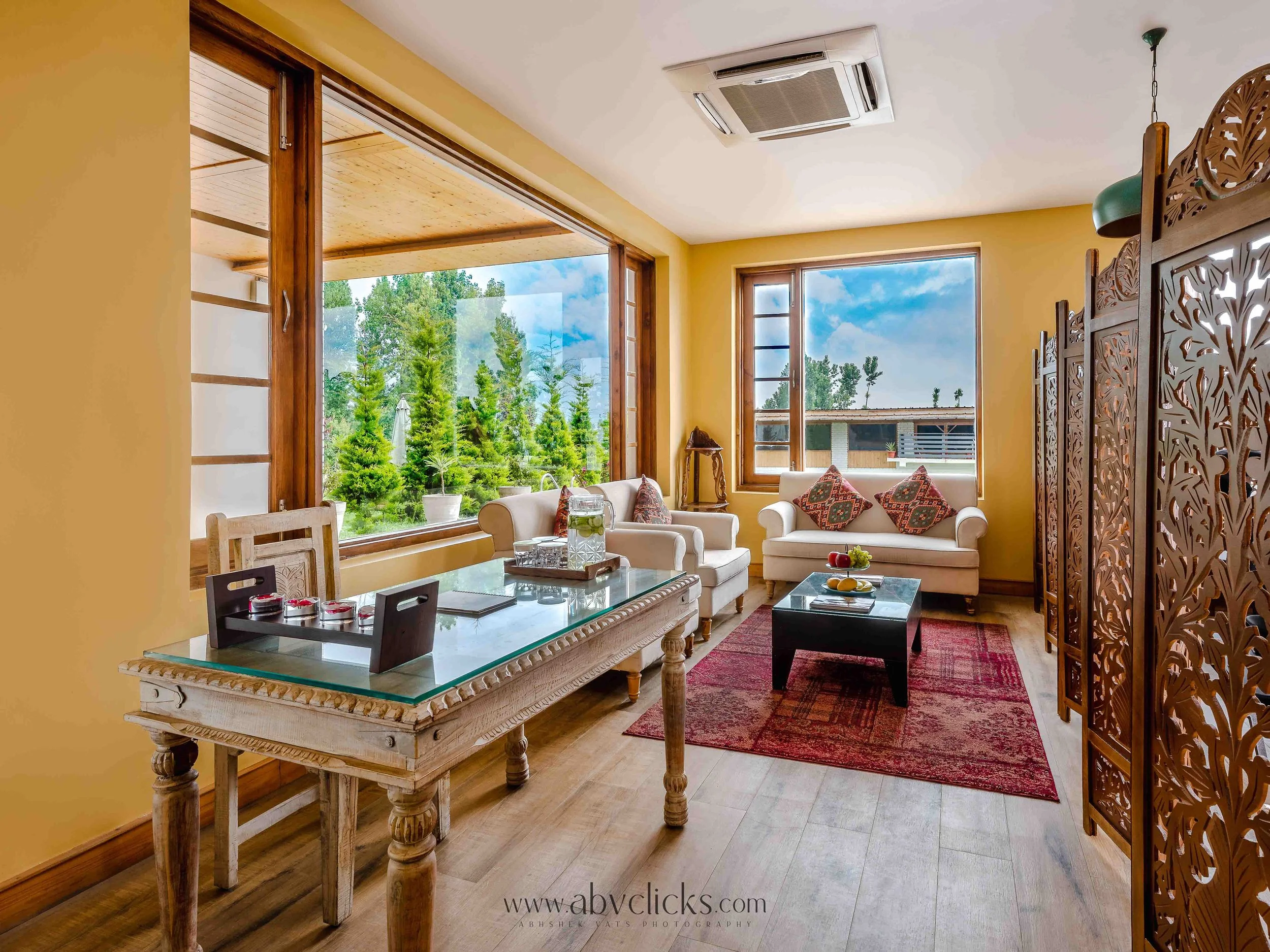 Living room with large windows showing greenery outside, white sofas with patterned pillows, a glass-topped wooden coffee table with fruit, and a decorative wooden room divider.