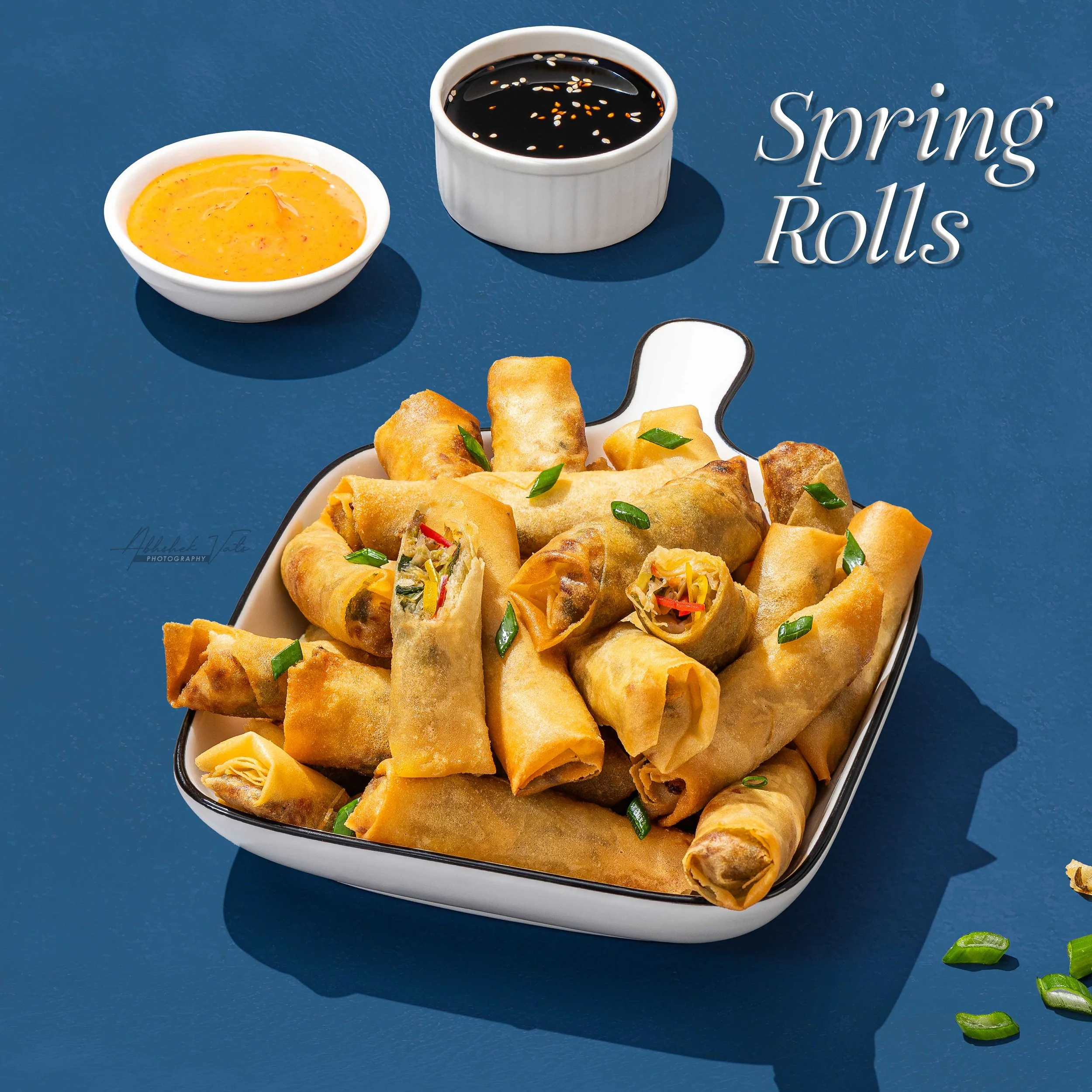 Plate of spring rolls garnished with green onions, accompanied by a small bowl of dipping sauce and a small bowl of orange-colored sauce with herbs, all on a blue surface.