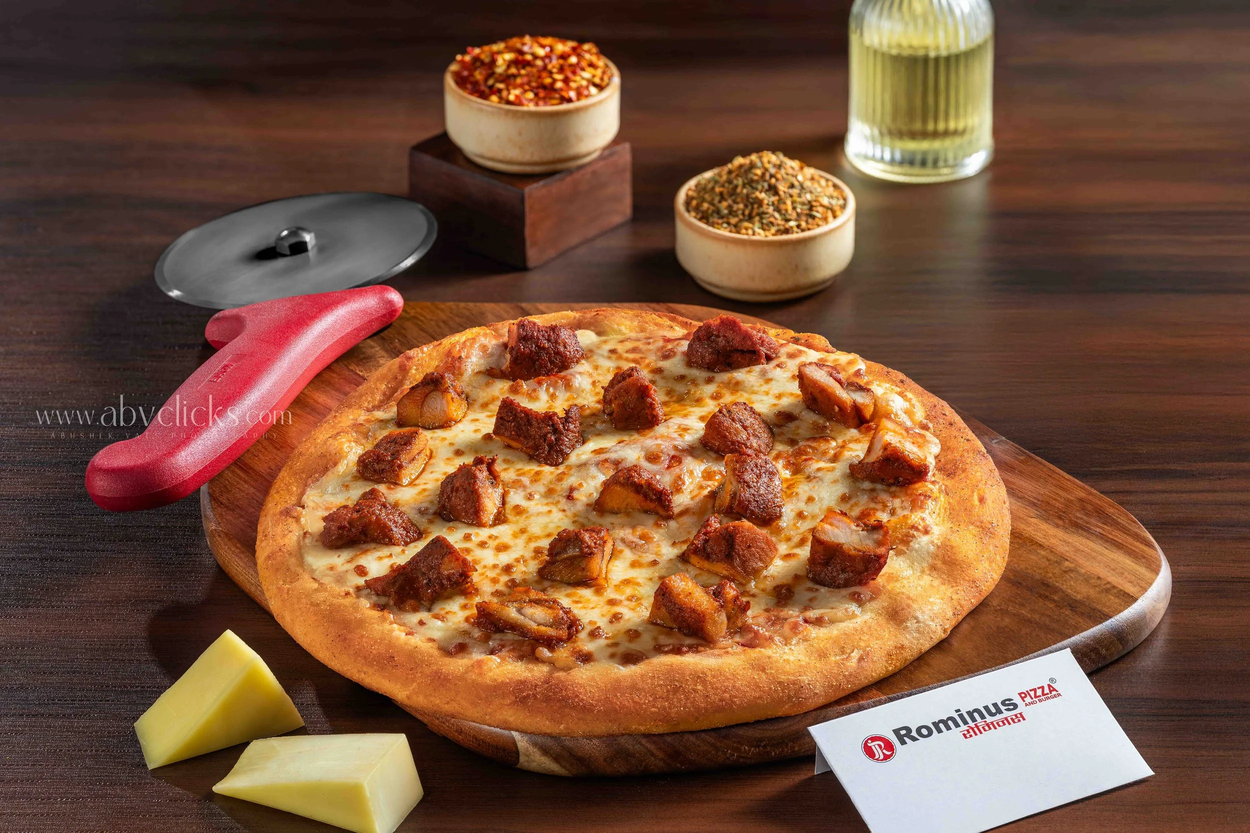 A cheese pizza with chunks of meat on a wooden board, next to a small wedge of cheese, with spice containers, a pizza cutter, a glass of water, and a menu printed on a card in the background.