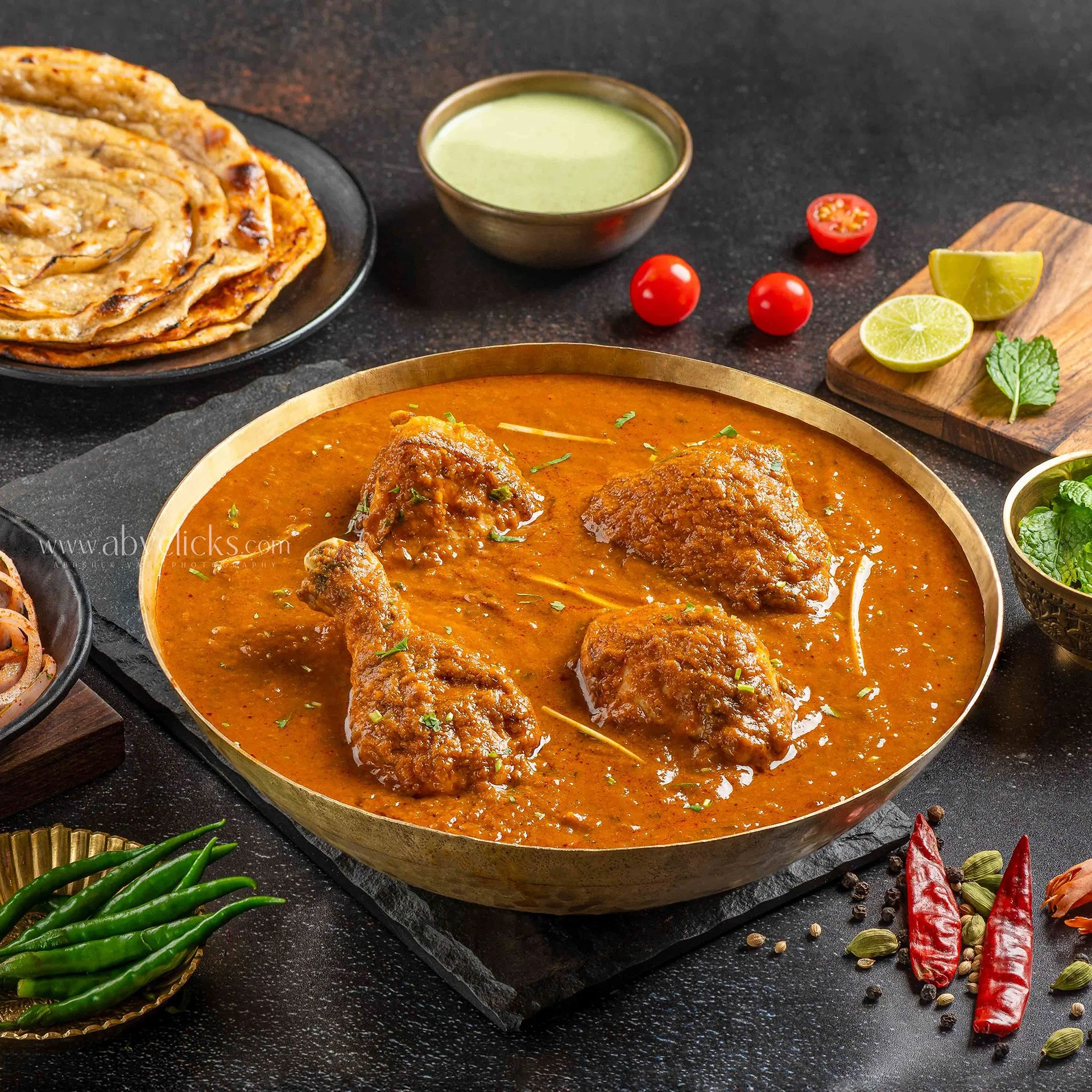 A close-up of a traditional Indian meal showing a bowl of butter chicken curry with chicken pieces in a creamy tomato-based sauce, green chilies, lime wedges, chili peppers, and a plate of layered flatbread, naan, on a dark textured surface.