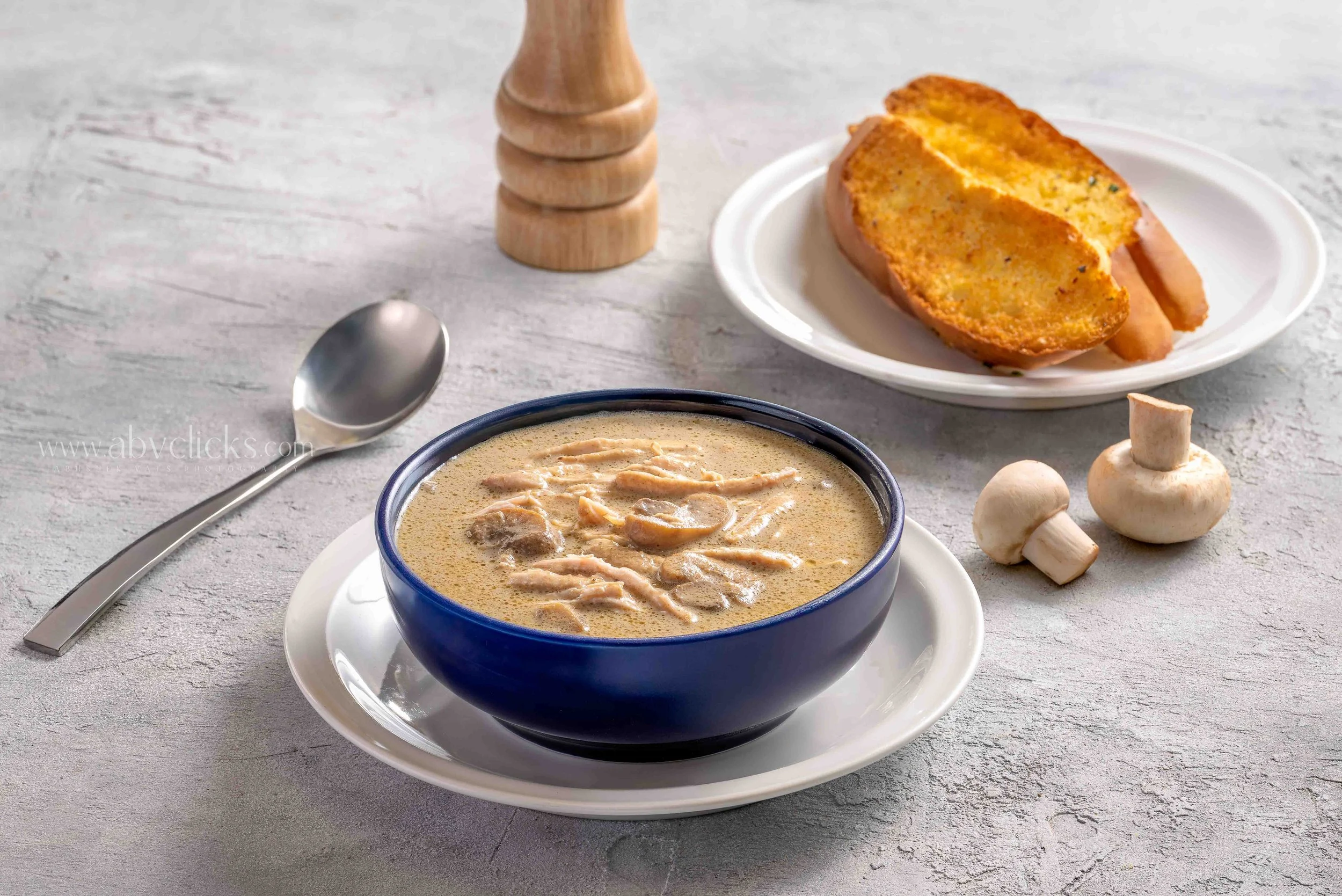 A bowl of creamy mushroom soup garnished with shredded chicken, placed on a white plate with a silver spoon beside it. In the background, there is a plate with a sliced toasted bread roll, several mushrooms, a wooden pepper grinder, and a light-color