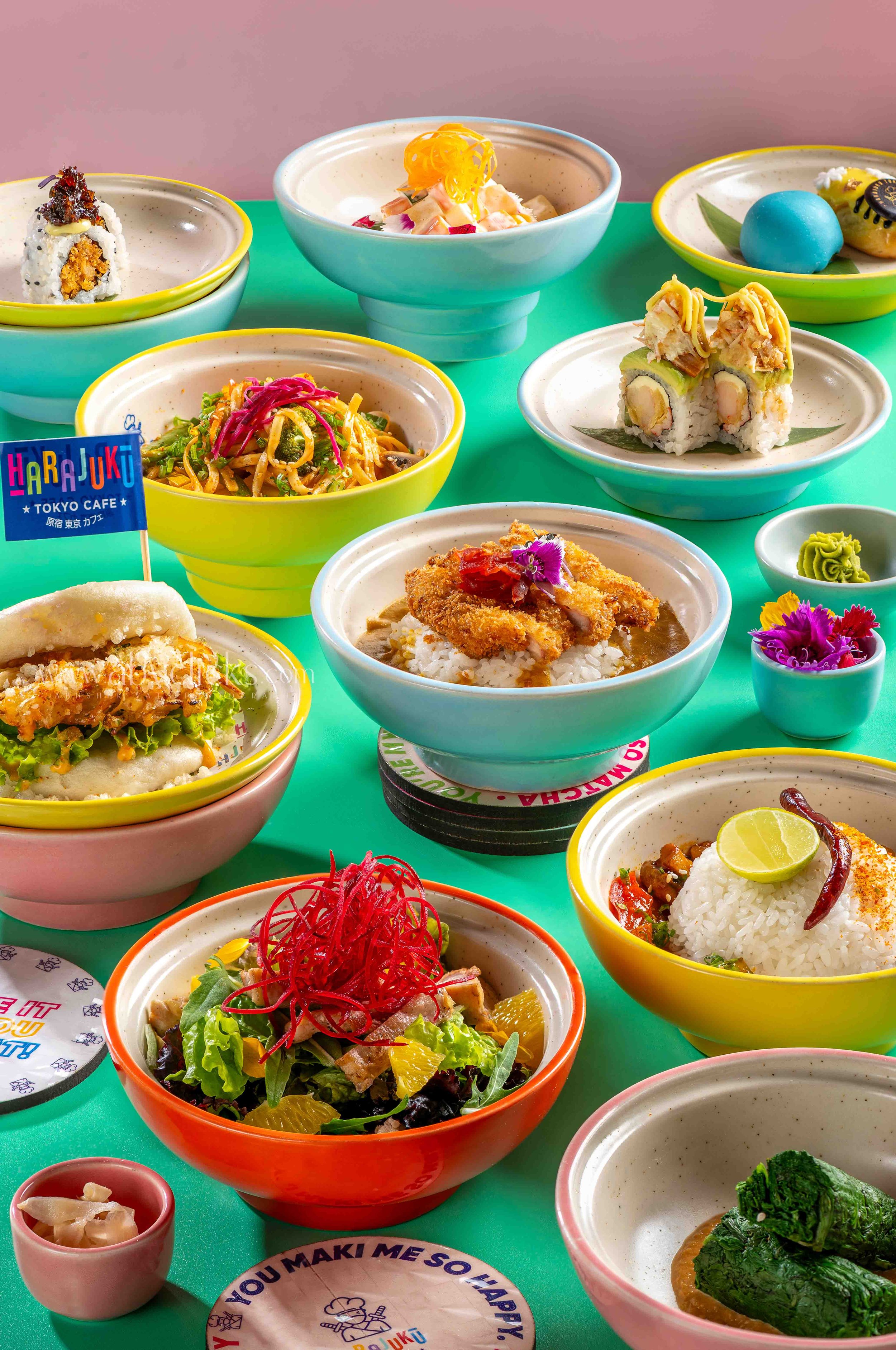 Colorful Japanese food dishes including sushi rolls, salads, rice bowls, and desserts, served on a green table with small dipping bowls.