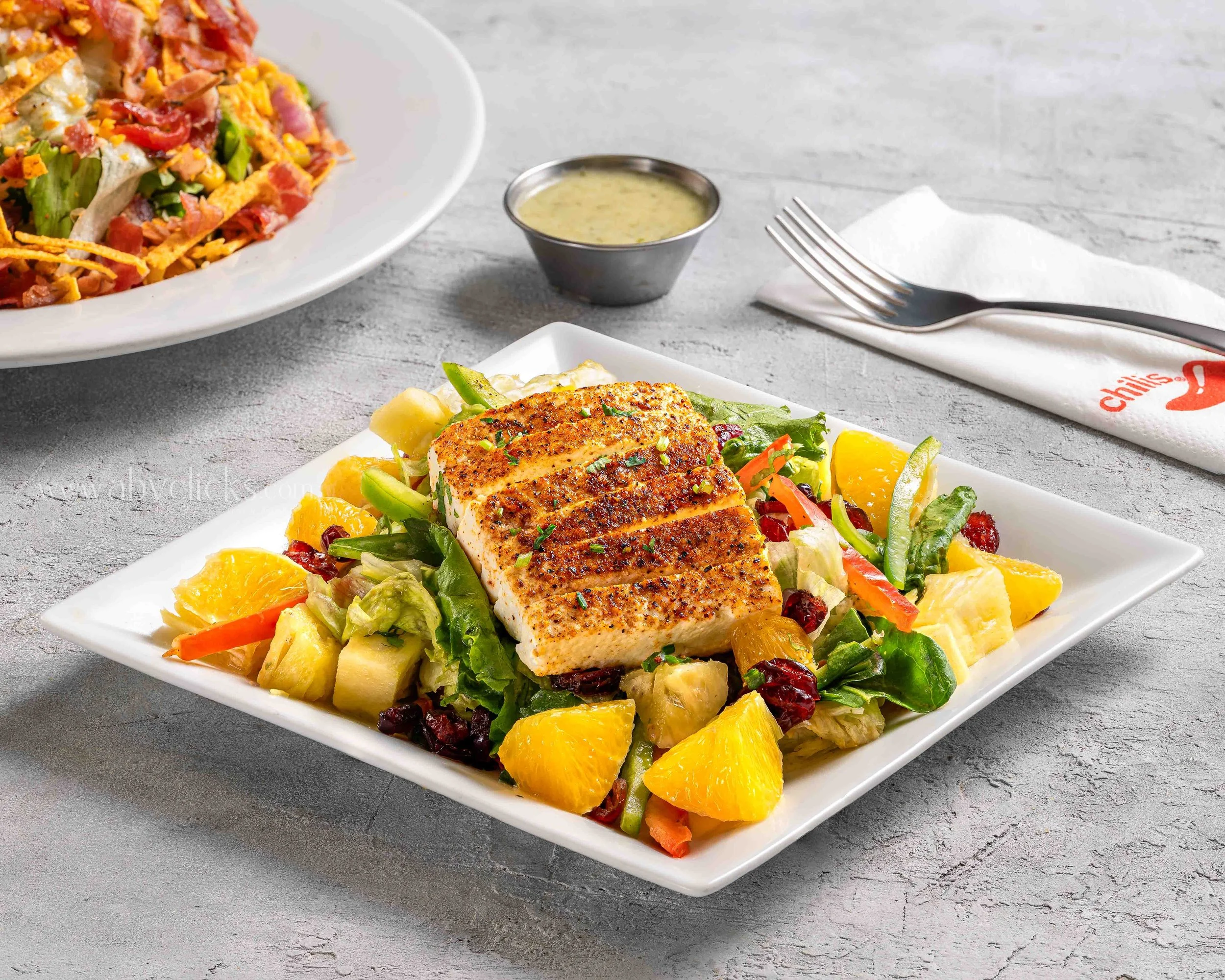 A white rectangular plate with seasoned pan-seared fish topped with chopped green herbs, served on a bed of mixed vegetables including lettuce, carrots, cauliflower, and orange segments. A small cup of yellowish sauce, a fork on a napkin, and another