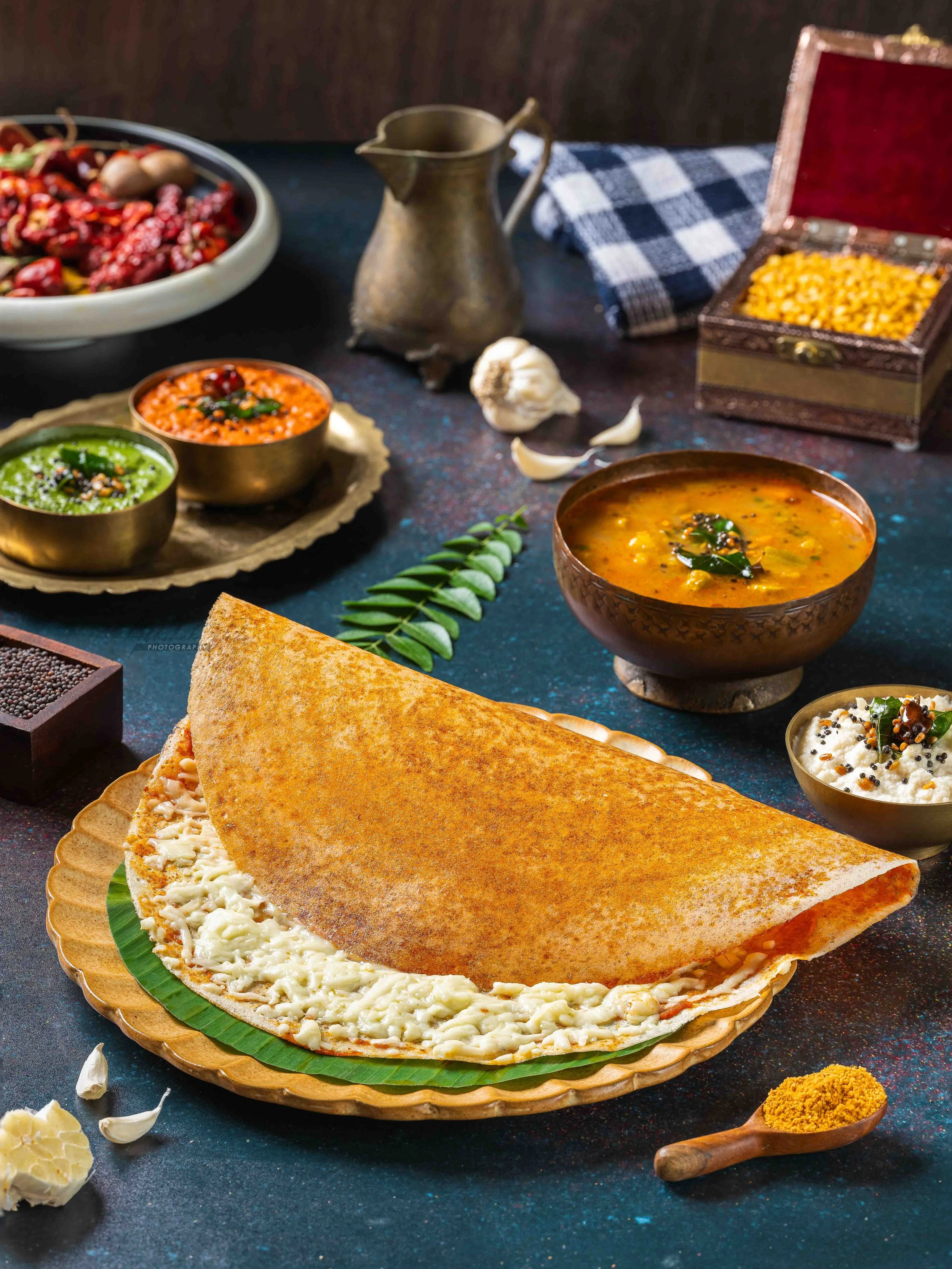 A plate of cheese-filled paratha on a wooden dish, accompanied by bowls of cooked vegetables and condiments, with garlic, spices, and a metal pitcher on a dark textured surface.