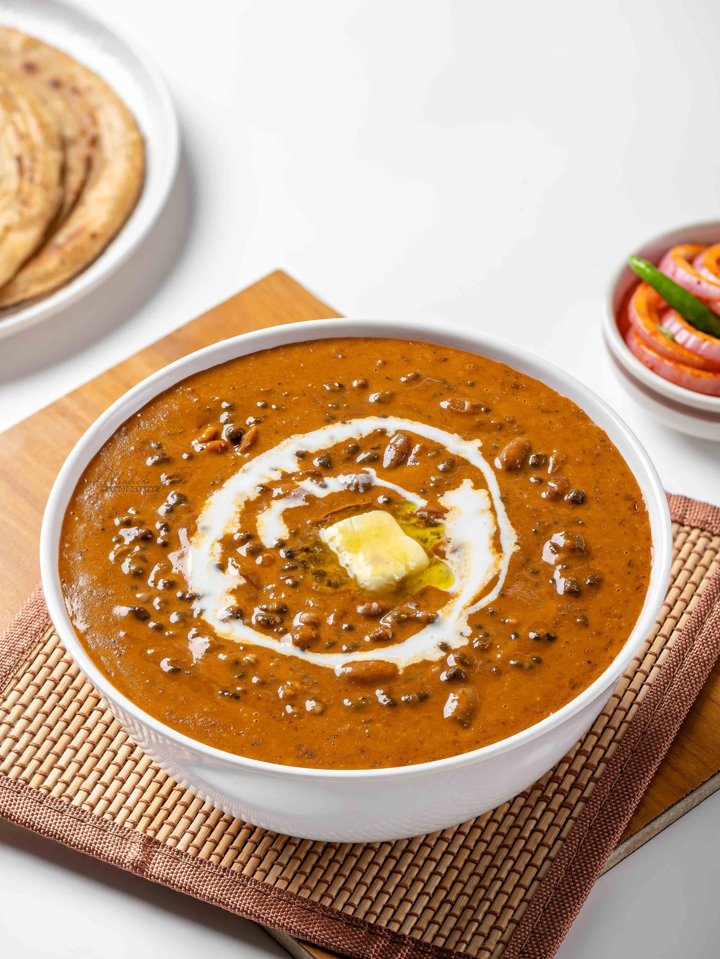 A bowl of Indian lentil soup garnished with cream and butter, placed on a woven placemat with a side of crispy bread and a small bowl of pickled vegetables.