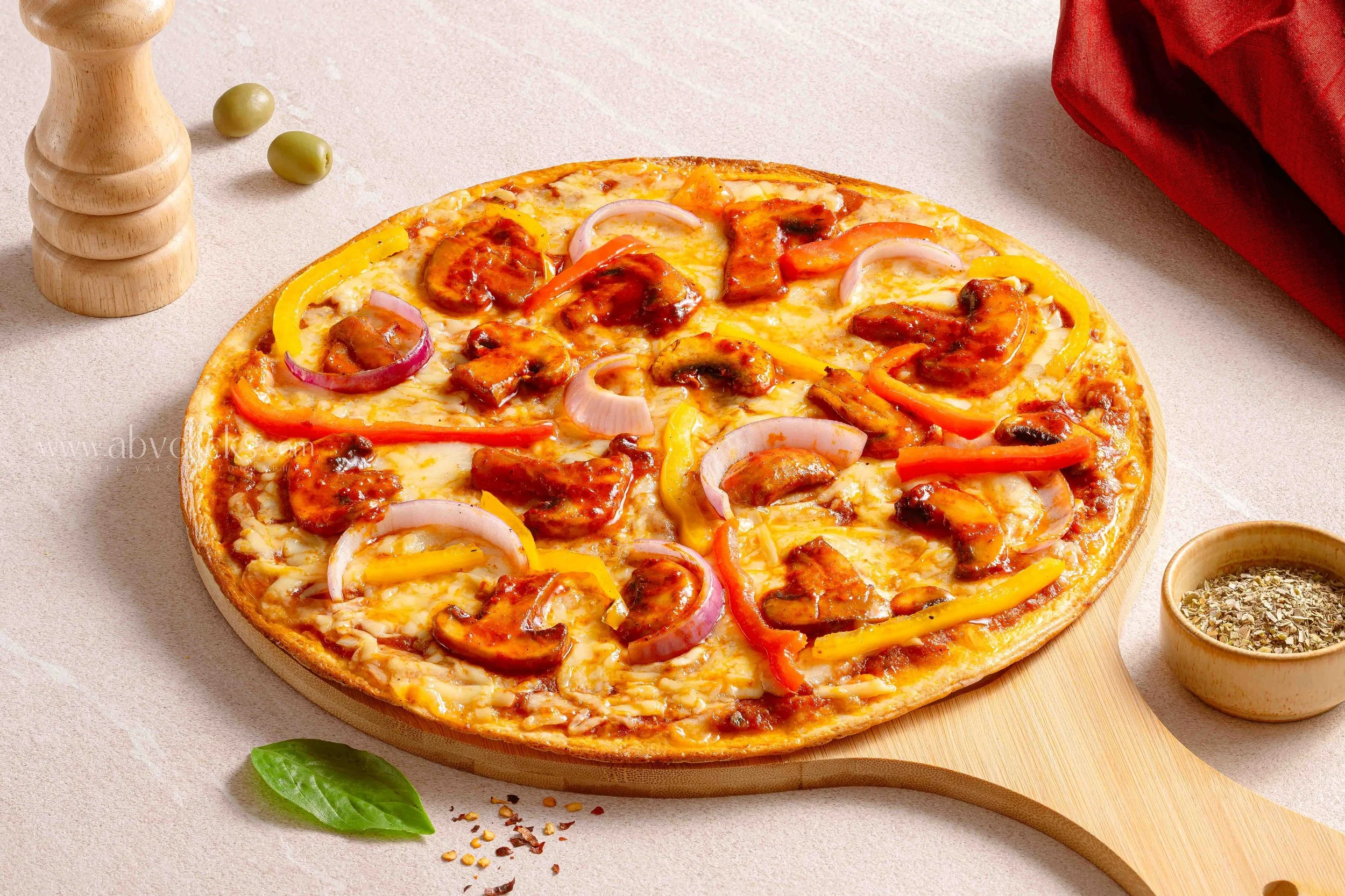 A pizza with cheese, red and yellow peppers, onions, and chicken on a wooden pizza peel, with a red napkin and bowls of herbs and spices nearby on a light-colored surface.