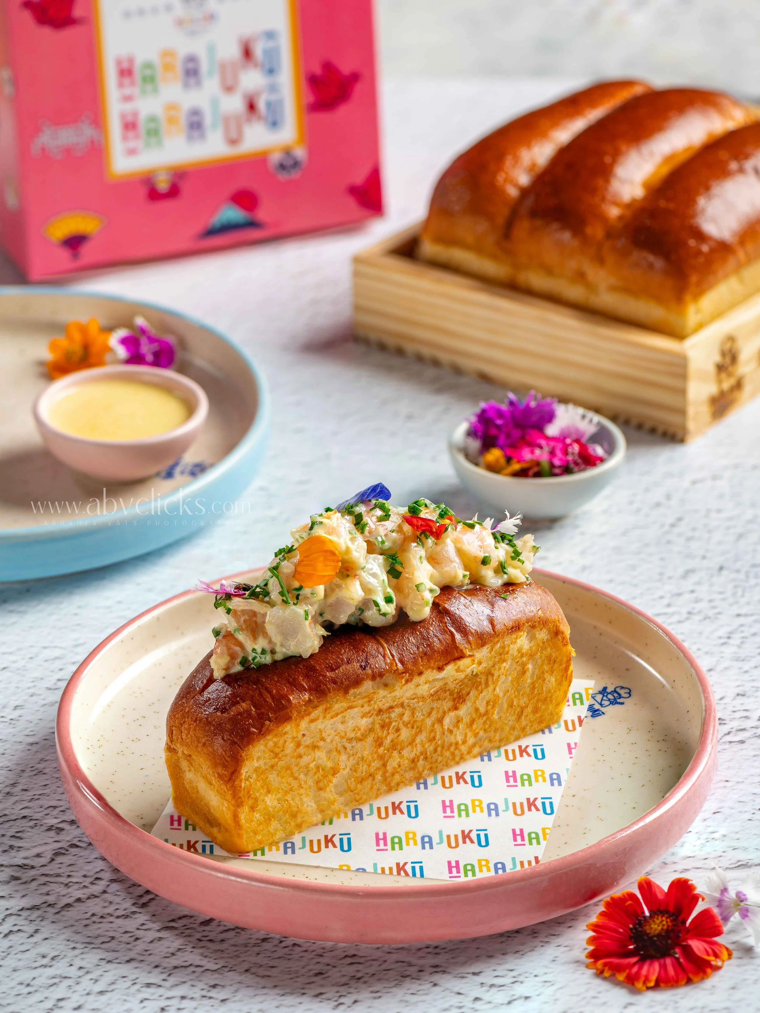 Close-up of a toasted bread roll topped with a creamy potato salad garnished with chopped chives and edible flowers on a pink plate with colorful 'Hajuku' paper liner.