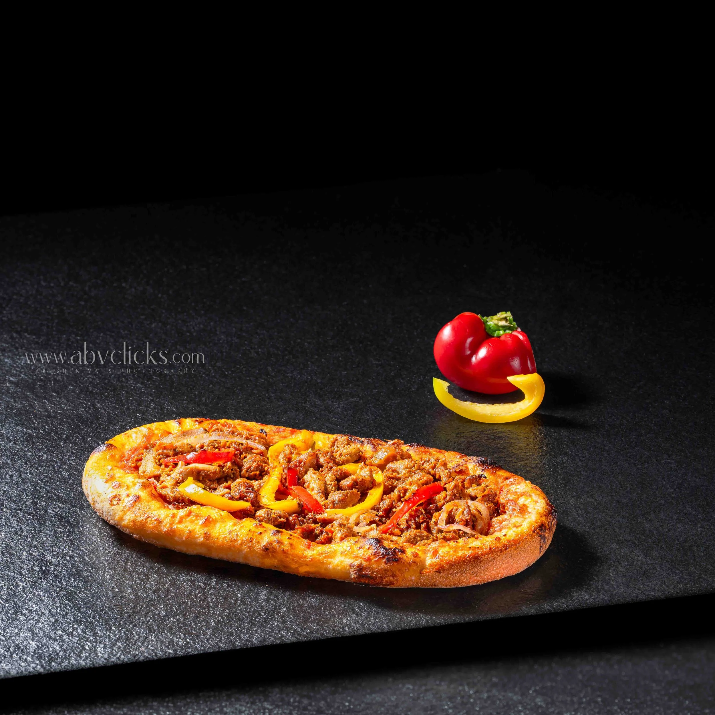 A slice of pizza topped with ground meat, red and yellow bell peppers, placed on a black surface with a small garnish of cherry tomatoes and bell pepper slices in the background.