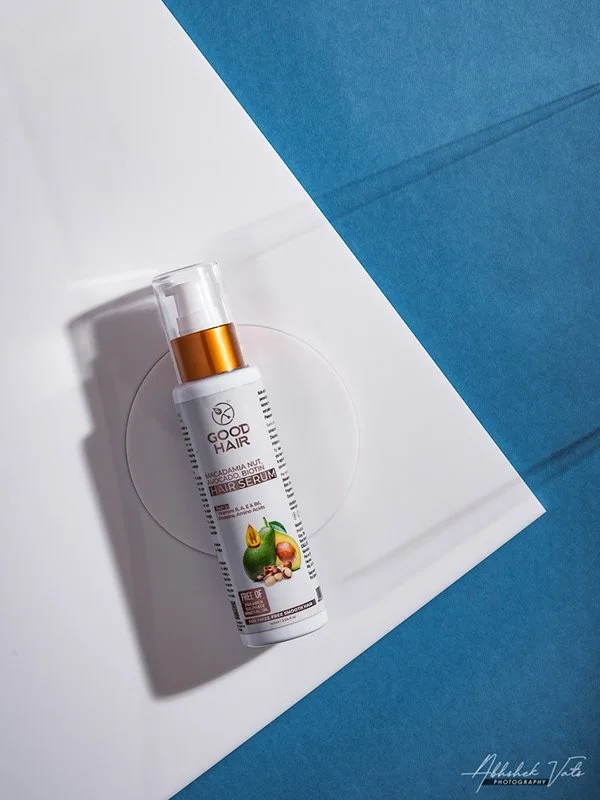 A white bottle of GOOD HAIR serum with a gold cap and pump, featuring avocado, nut, and fig illustrations, placed on a white circular platform with blue and white papers underneath.
