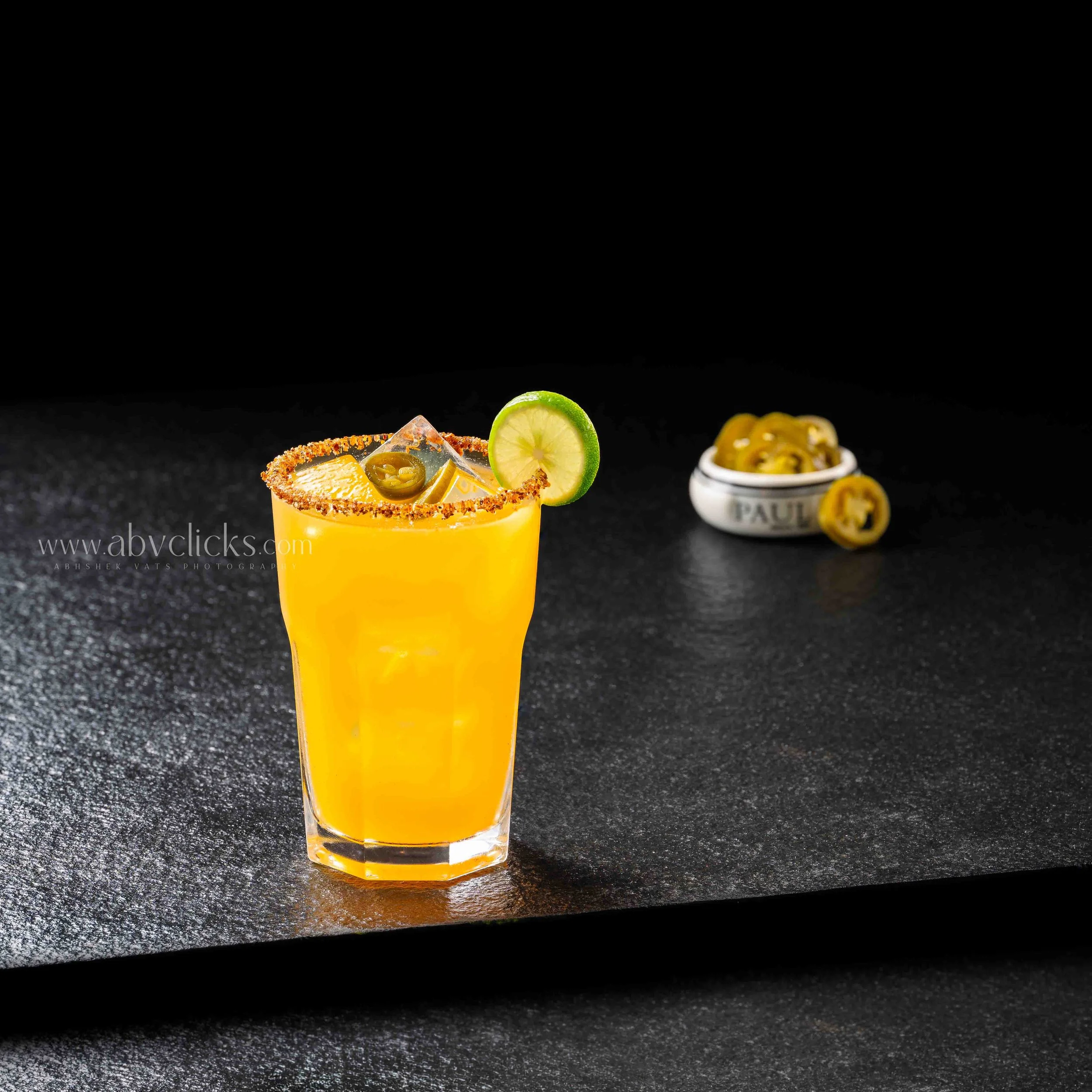 Close-up of a yellow cocktail in a tall glass garnished with a lime wedge and jalapeño slices, on a black surface with a black background.