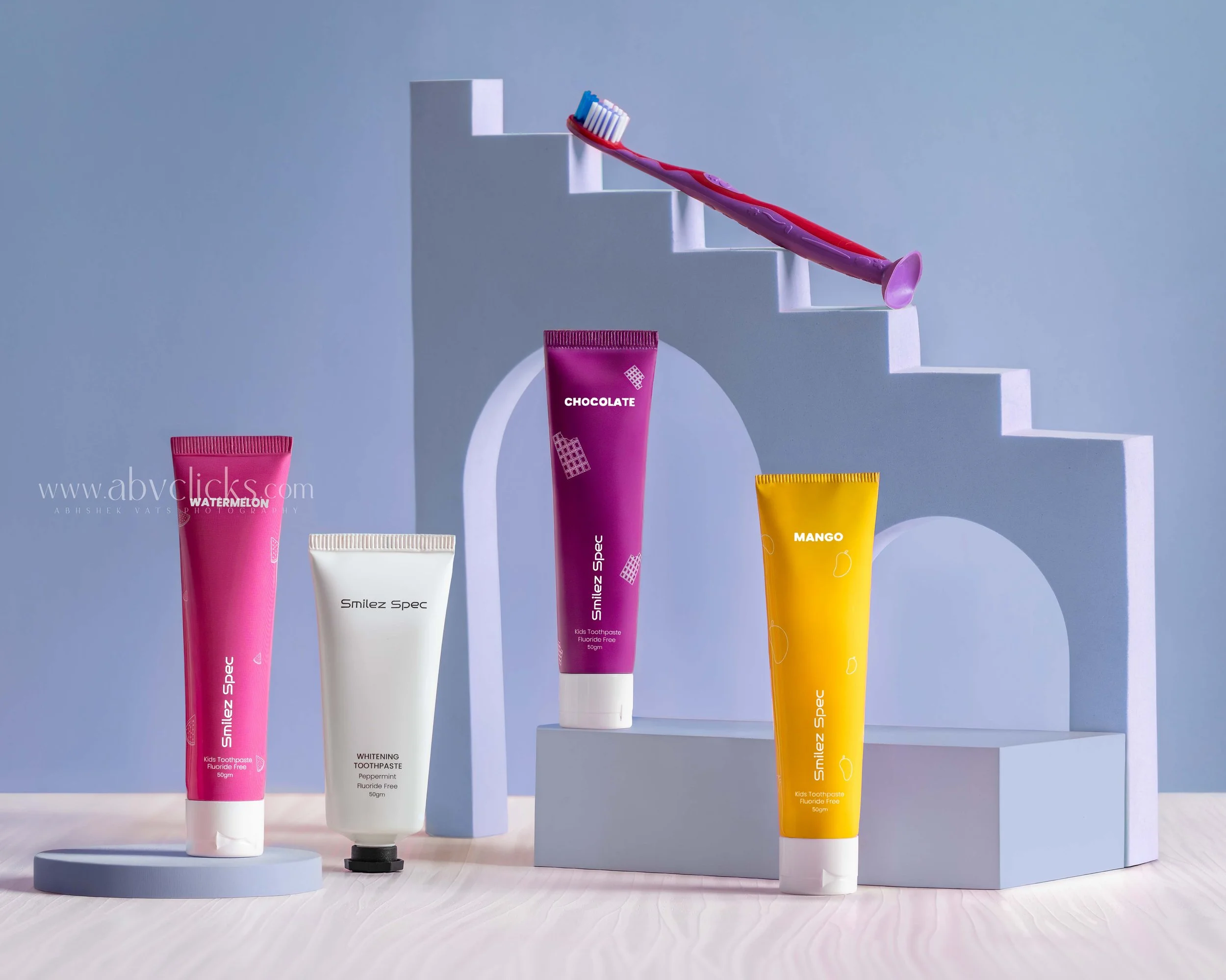 Assorted tubes of children's toothpaste in pink, white, purple, and yellow on display stands, with a toothbrush resting on one of the tubes.