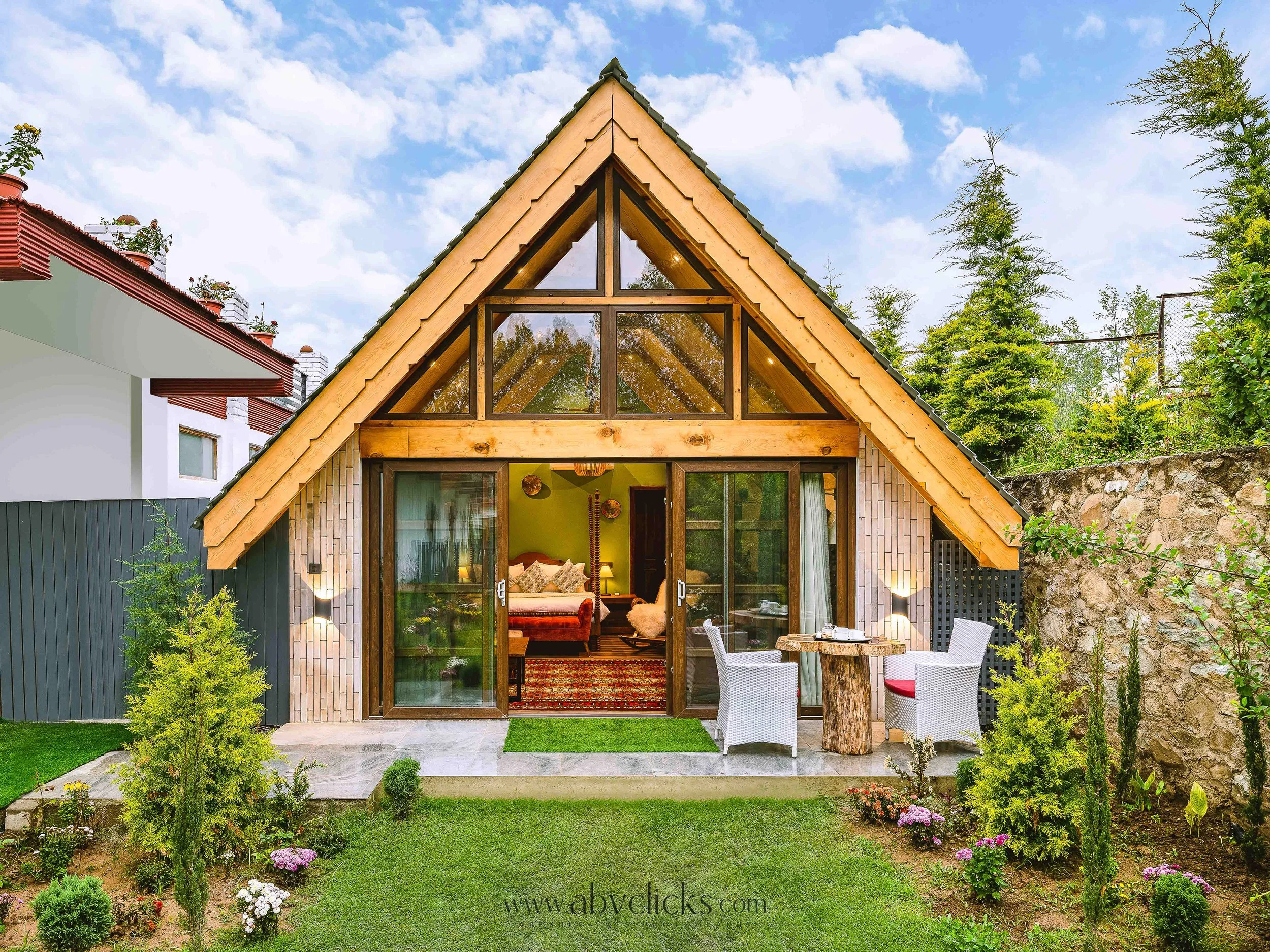 A modern wooden A-frame house with a large triangular front window and sliding glass doors opening to a garden with outdoor furniture, surrounded by green trees and plants under a cloudy sky.
