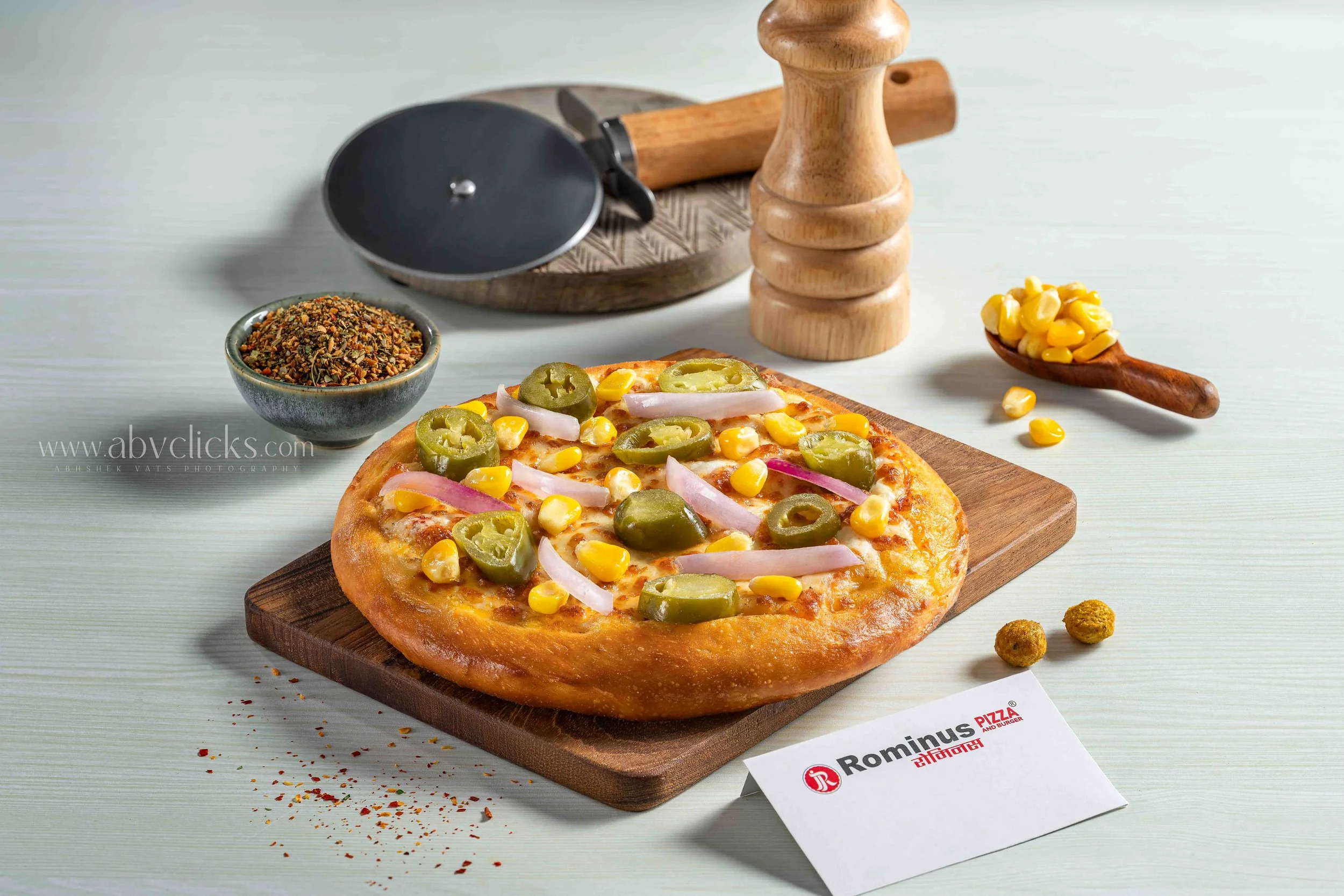 A pizza topped with jalapeños, onions, and corn on a wooden cutting board. Nearby are a bowl of spices, a black pizza cutter, a wooden pepper grinder, and a handful of corn kernels on a spoon. A Romano's pizza receipt is partially visible in the fore