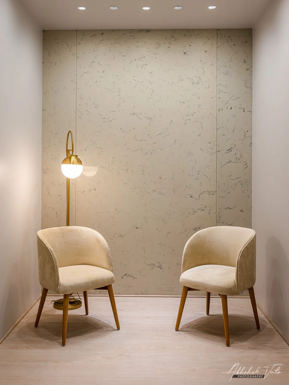Two beige upholstered armchairs with wooden legs in a small, minimalist room with a marble wall, a floor lamp with a round white shade, and ceiling recessed lights.