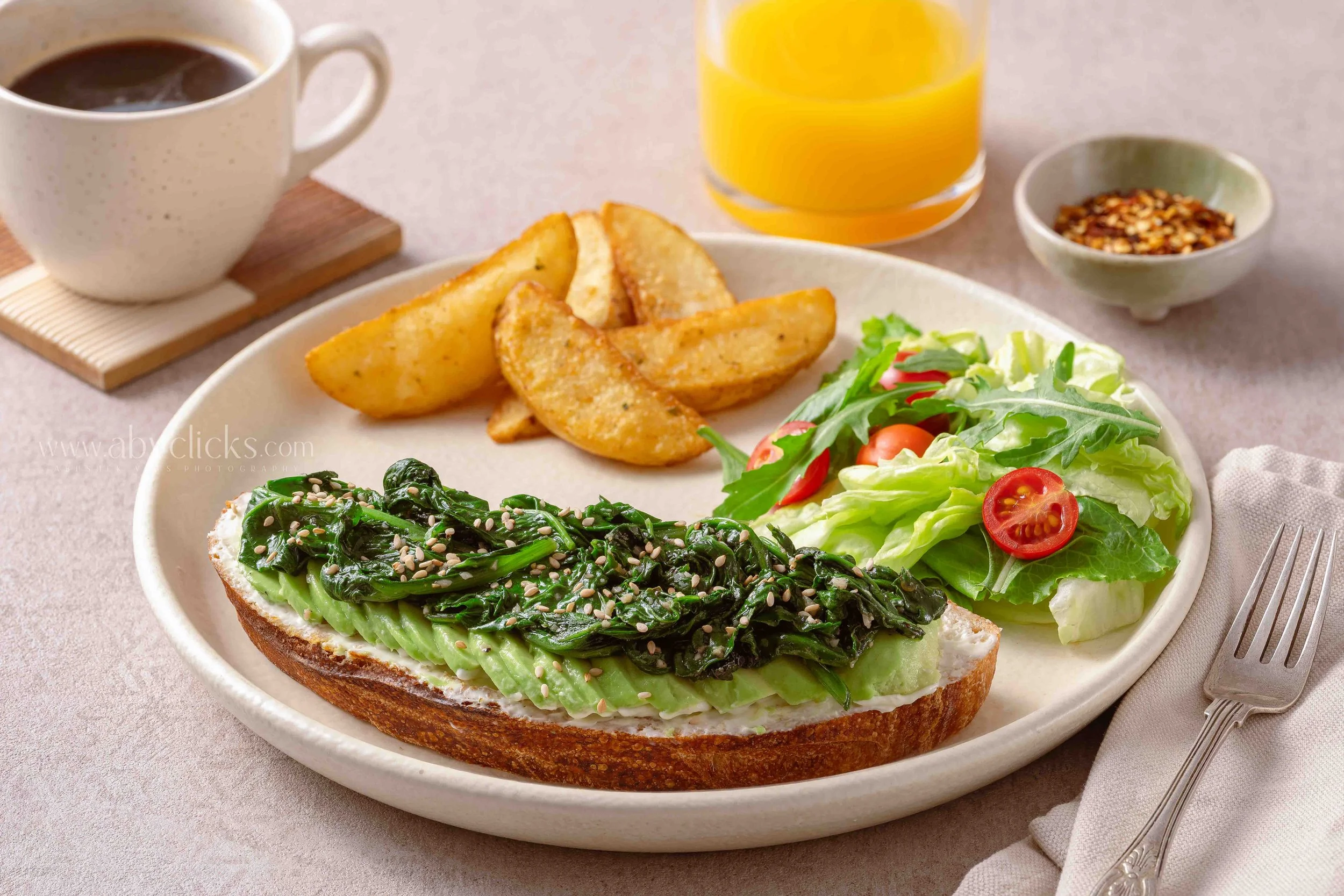 A breakfast plate with toast topped with avocado and greens, side salad with cherry tomatoes, potato wedges, a glass of orange juice, a cup of black coffee, and a small bowl of red pepper flakes.