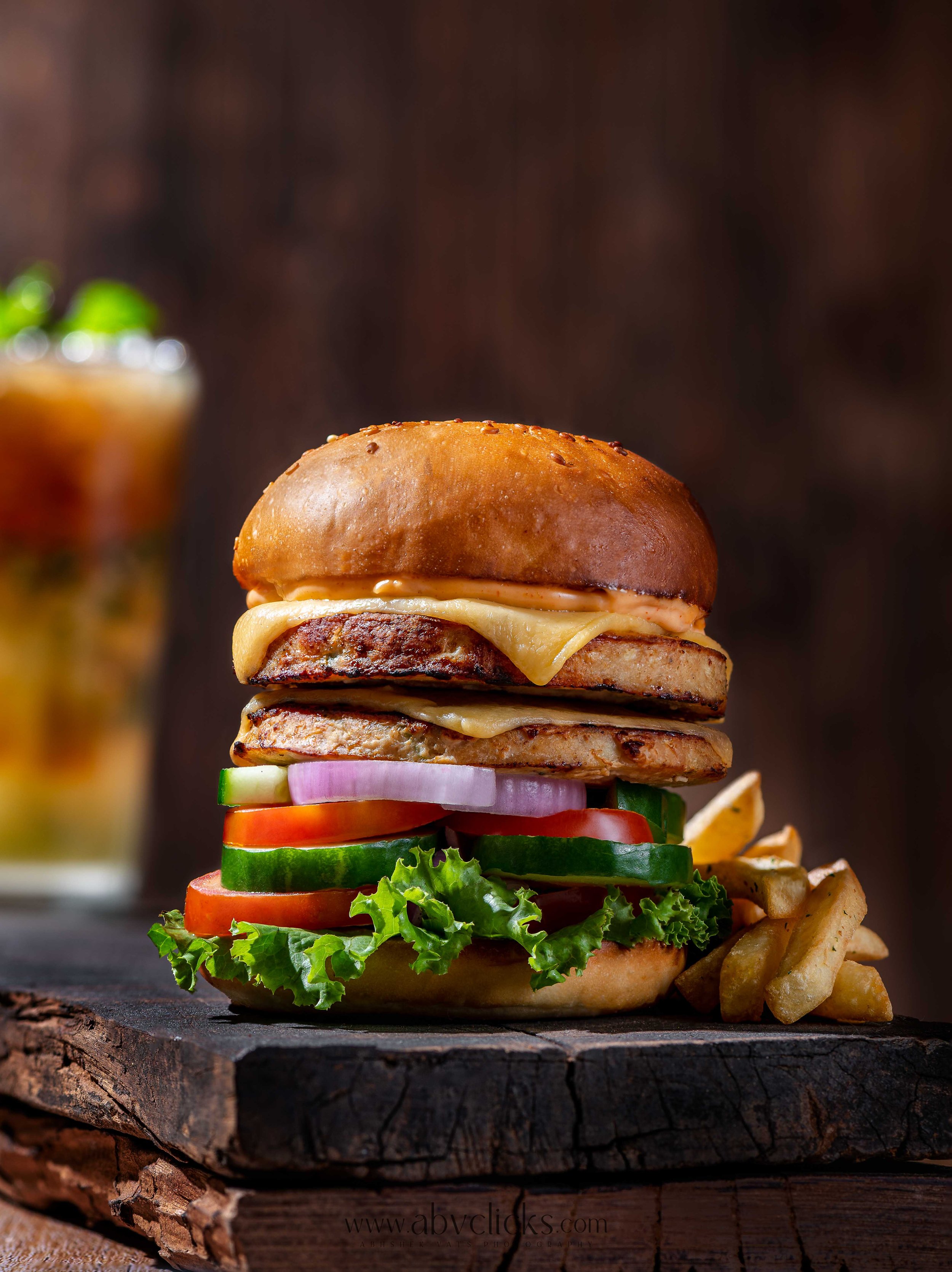 A tall burger with multiple layers of lettuce, tomato, cucumber, onion, cheese, beef patties, and a toasted bun, plated on a wooden surface with a side of fries.