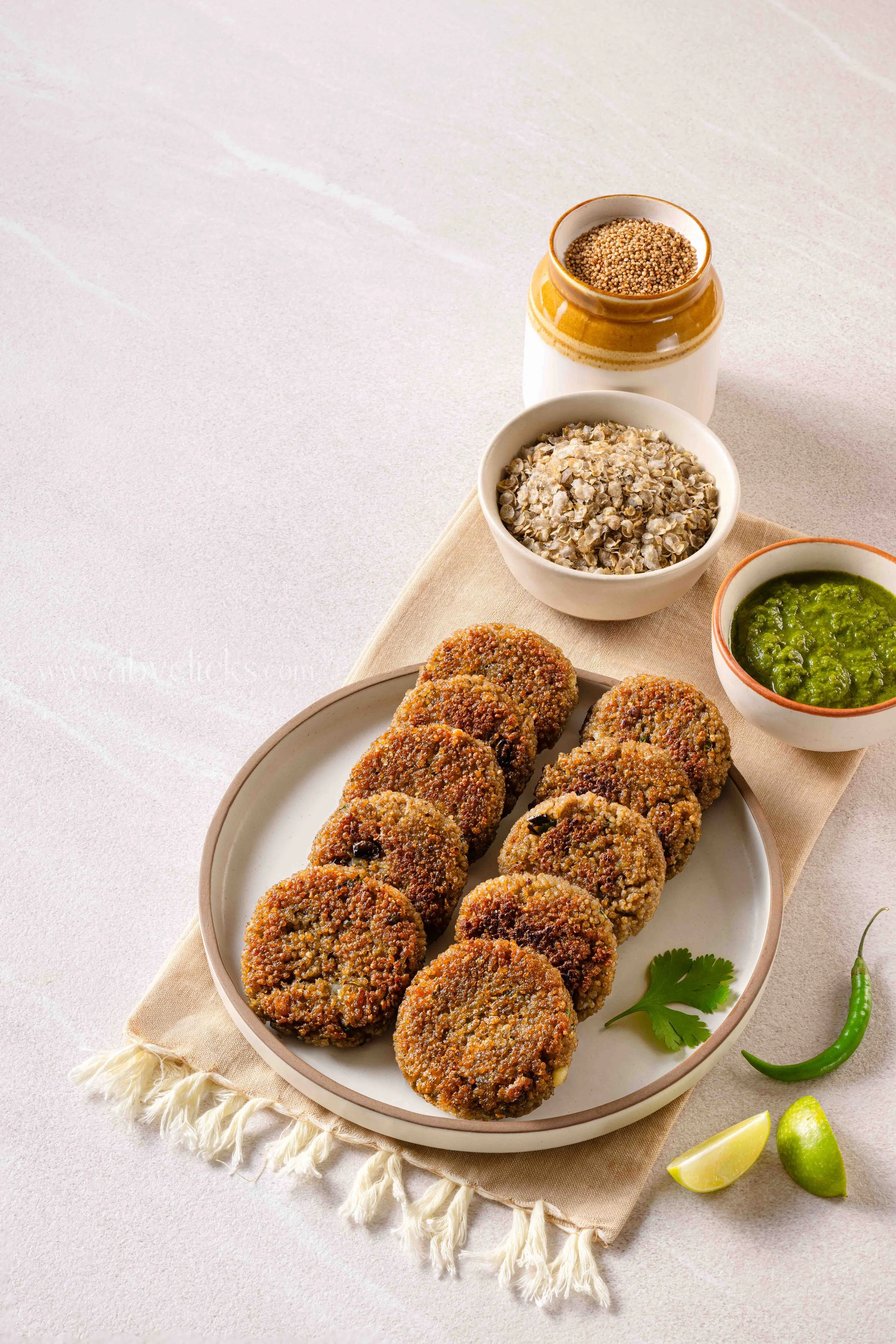 Plate of falafel with green chili sauce, accompanied by lime wedges, cilantro, grains, and pickled vegetables on a textured white surface.
