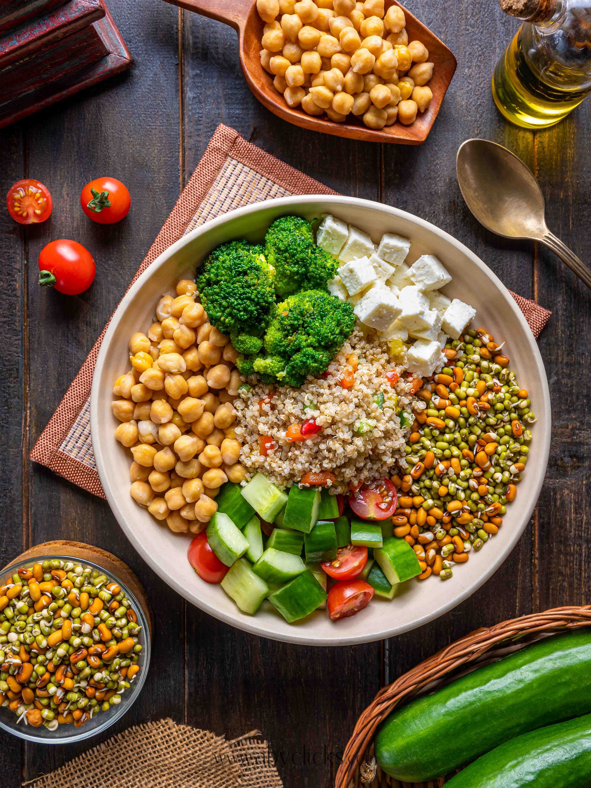 A large bowl of various fresh vegetables including cherry tomatoes, cucumber, and broccoli, with chickpeas, quinoa, and cubed feta cheese on top, surrounded by cherry tomatoes, chickpeas, a bottle of olive oil, zucchini, and a small bowl of sprouted 