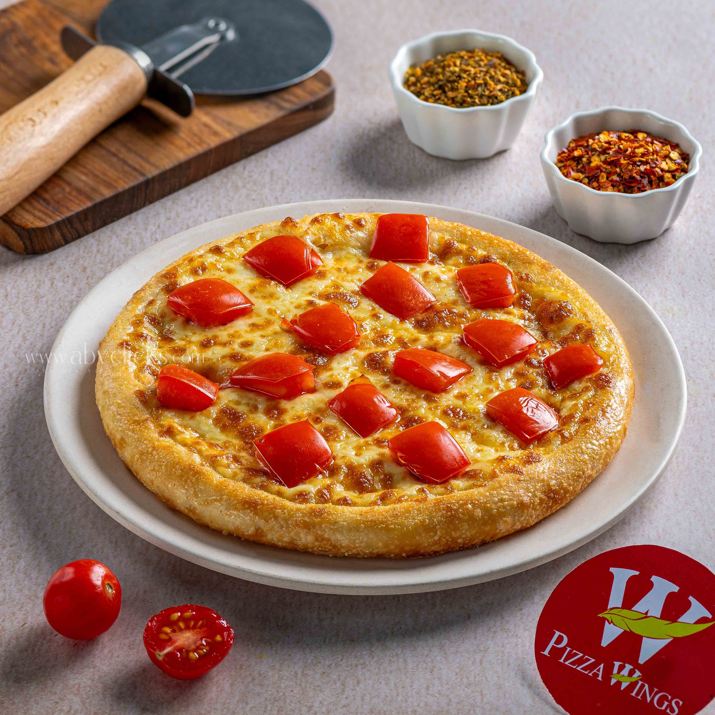 Close-up of a cheese and pepperoni pizza on a white plate, with cherry tomatoes and pizza box in the background, on a light-colored countertop.