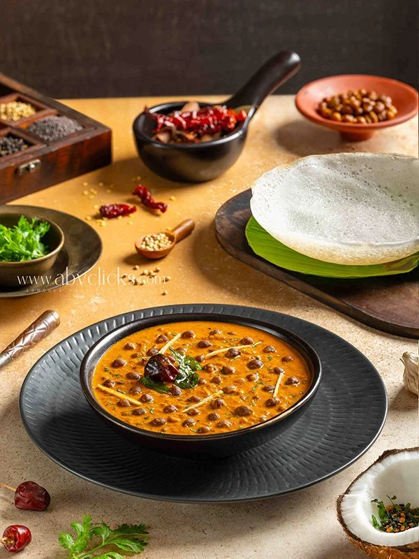 A bowl of Indian lentil soup garnished with herbs, placed on a decorative black plate, surrounded by various spices and ingredients including a green leafy vegetable, dried red chilies, sesame seeds, and a stack of rice paper sheets.
