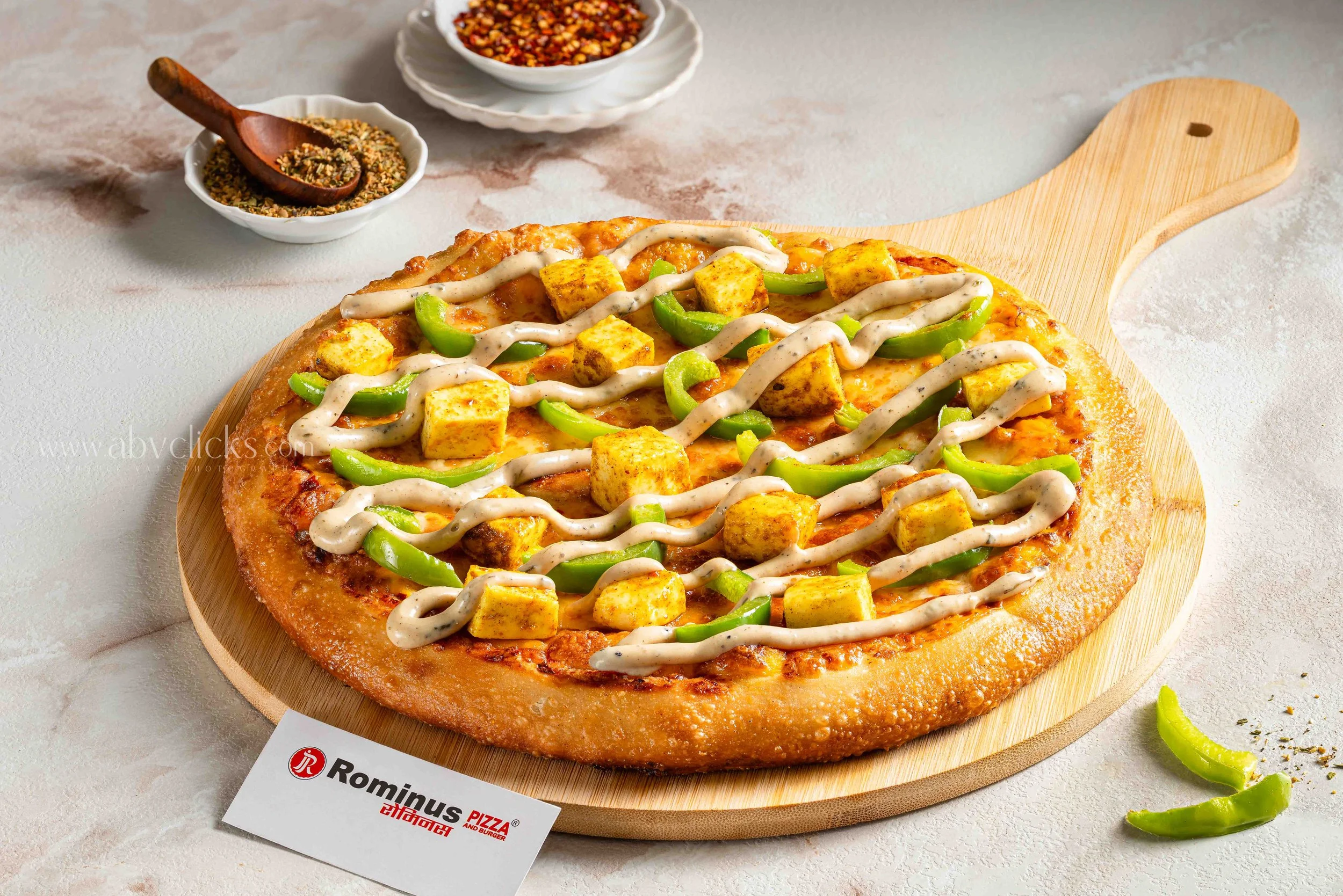 Pizza topped with green bell peppers and grilled chicken chunks on a wooden pizza board, with bowls of crushed red pepper flakes and herbs in the background.