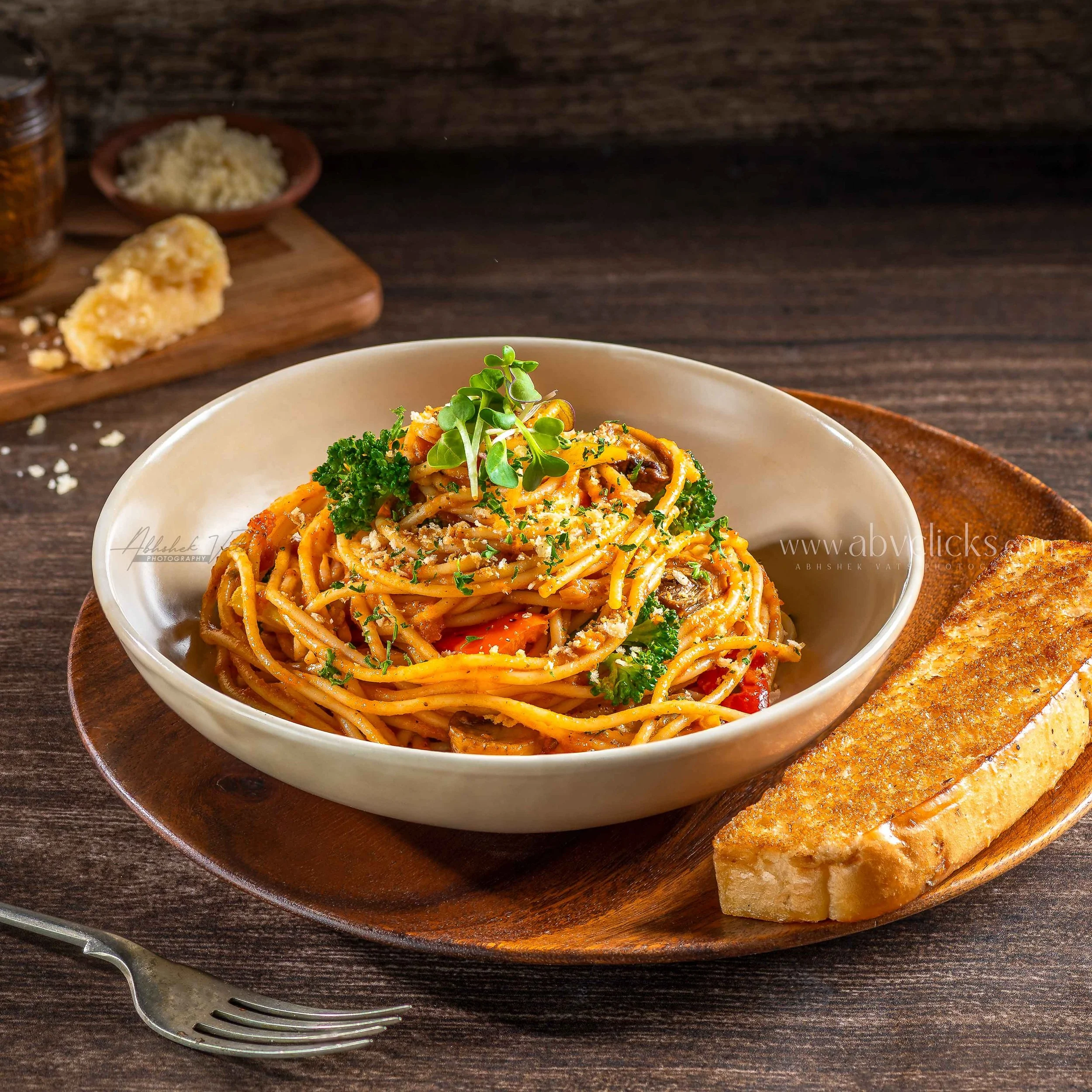 A bowl of spaghetti topped with herbs and grated cheese on a wooden plate, with bread and a fork in the background.