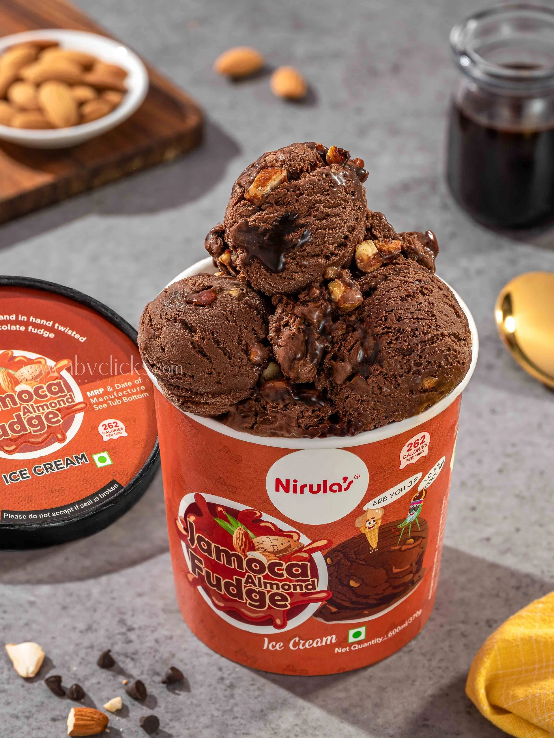 A container of Nirula's chocolate almond fudge ice cream with scoops on top, and a lid nearby. Scattered almonds, chocolate chips, and chopped nuts are on a gray surface, with a yellow cloth and a jar of syrup in the background.