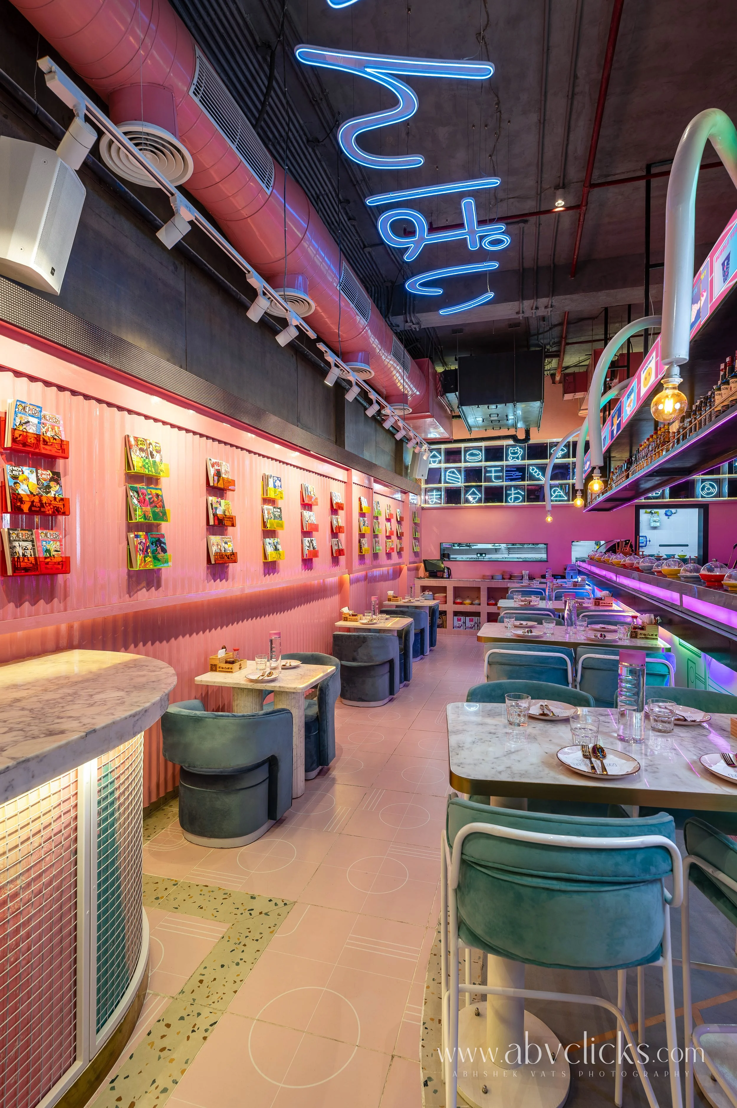 Brightly lit cafe interior with pink walls, marble tabletops, velvet chairs, and colorful neon signs on the wall and ceiling.