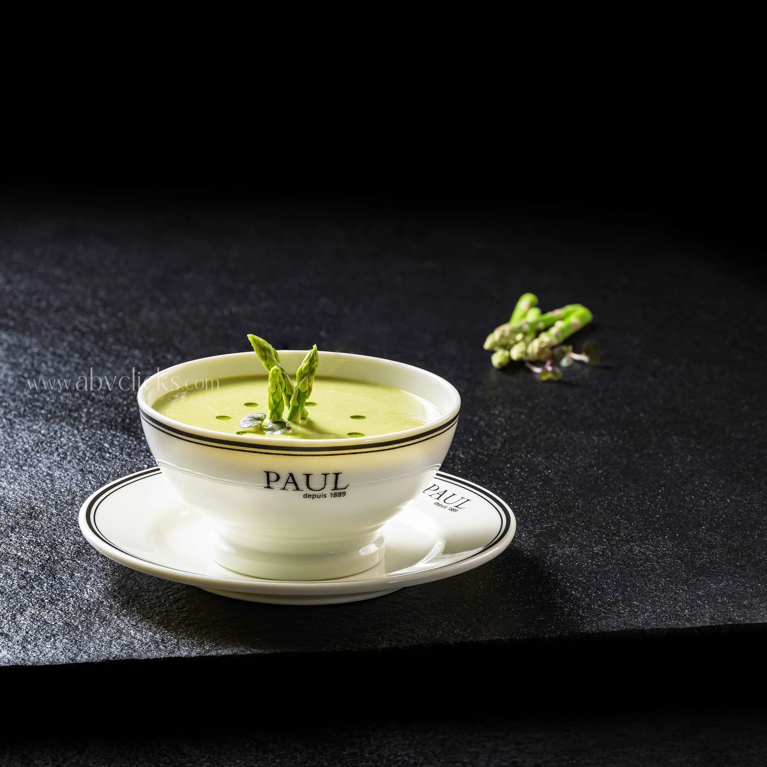 A small bowl with the word 'PAUL' on it, filled with green asparagus tips, with a scattering of some torn greens. The bowl is on a matching saucer on a black textured surface, with a plain black background.