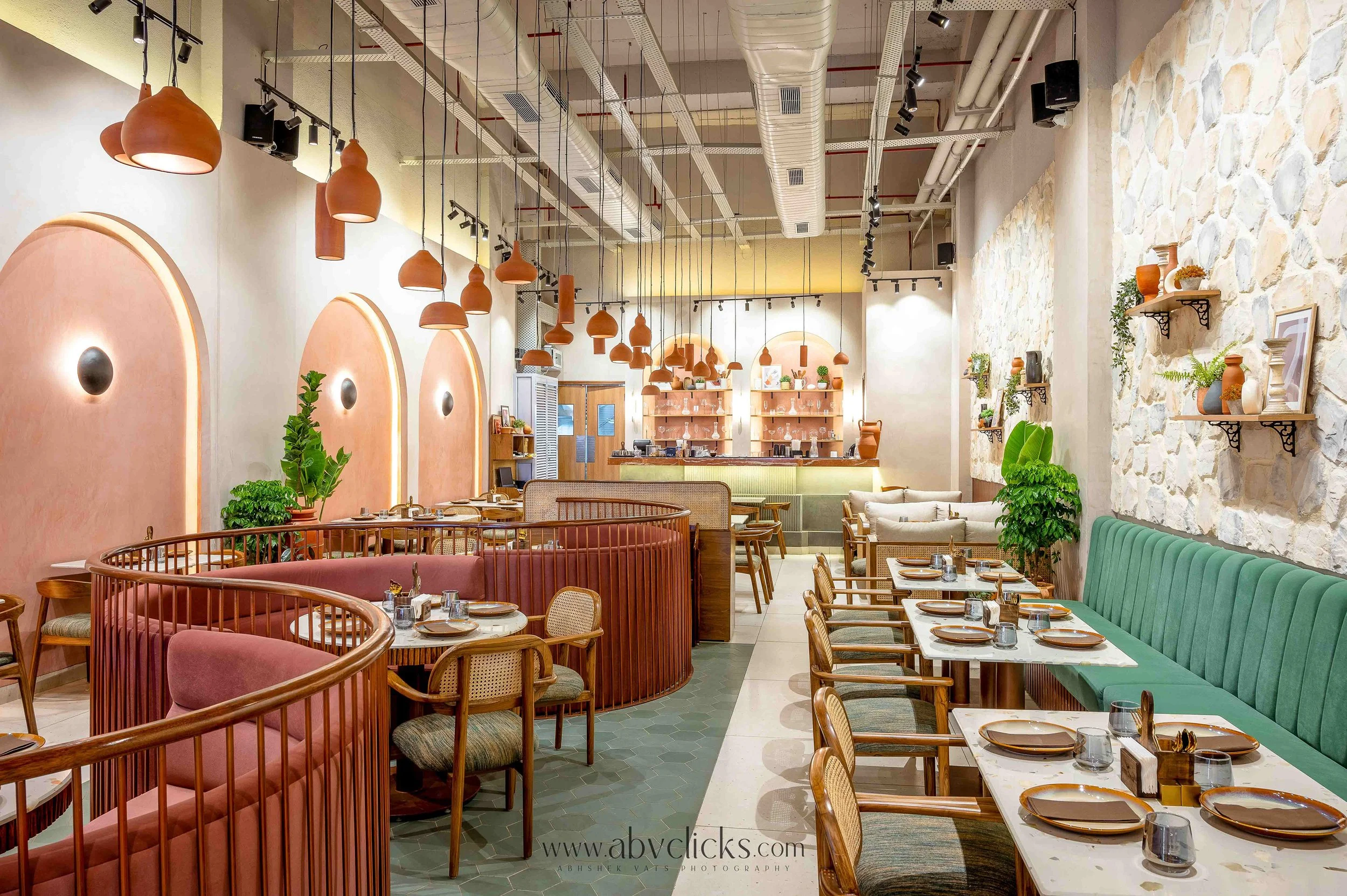 Interior of a restaurant with modern decor, terracotta pendant lights, a textured stone wall on the right, various green plants, and tables set with plates, glasses, and cutlery.