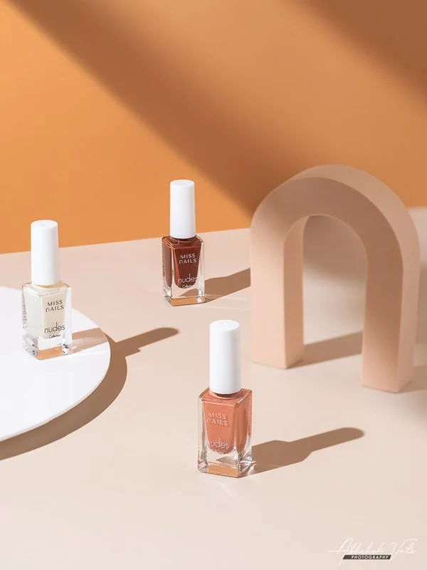 Three bottles of Miss Nails nail polish, two in nude shades and one in white, on a beige surface with a curved white platform and a pink arch-shaped object in the background, casting shadows.