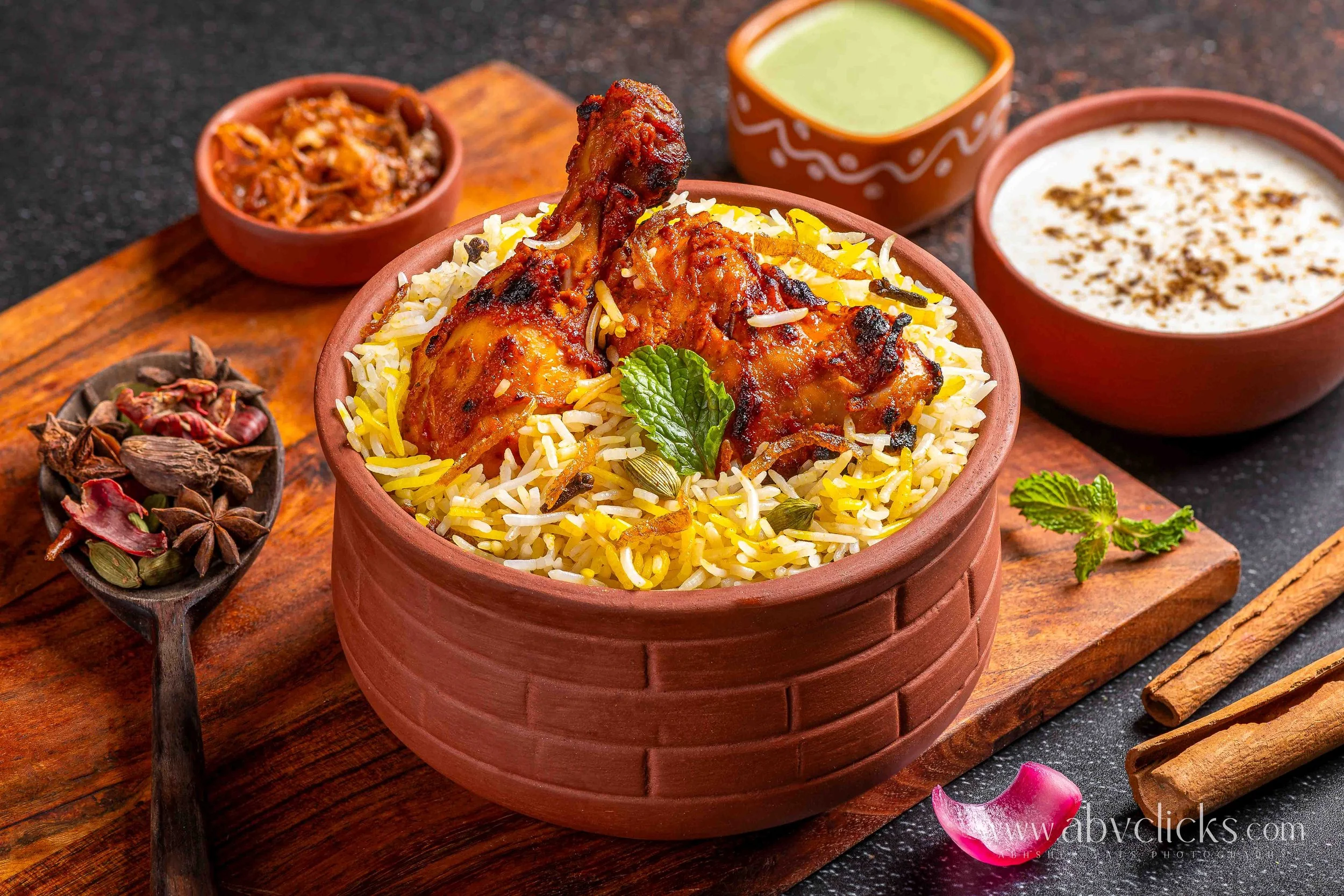 Close-up of a bowl of spiced chicken biryani garnished with fresh mint leaves, surrounded by small bowls of raita, curry, and spices on a wooden tray.