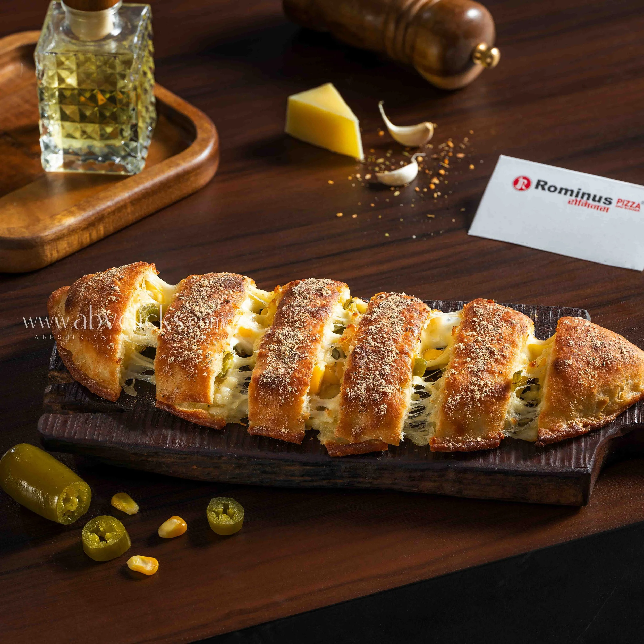 Close-up of a plate of cheese bread with a golden crust, sprinkled with cheese, on a dark wooden table. Next to the plate are slices of jalapeño peppers with seeds, a small piece of cheese, garlic cloves, a wooden pepper grinder, a bottle of oil on a