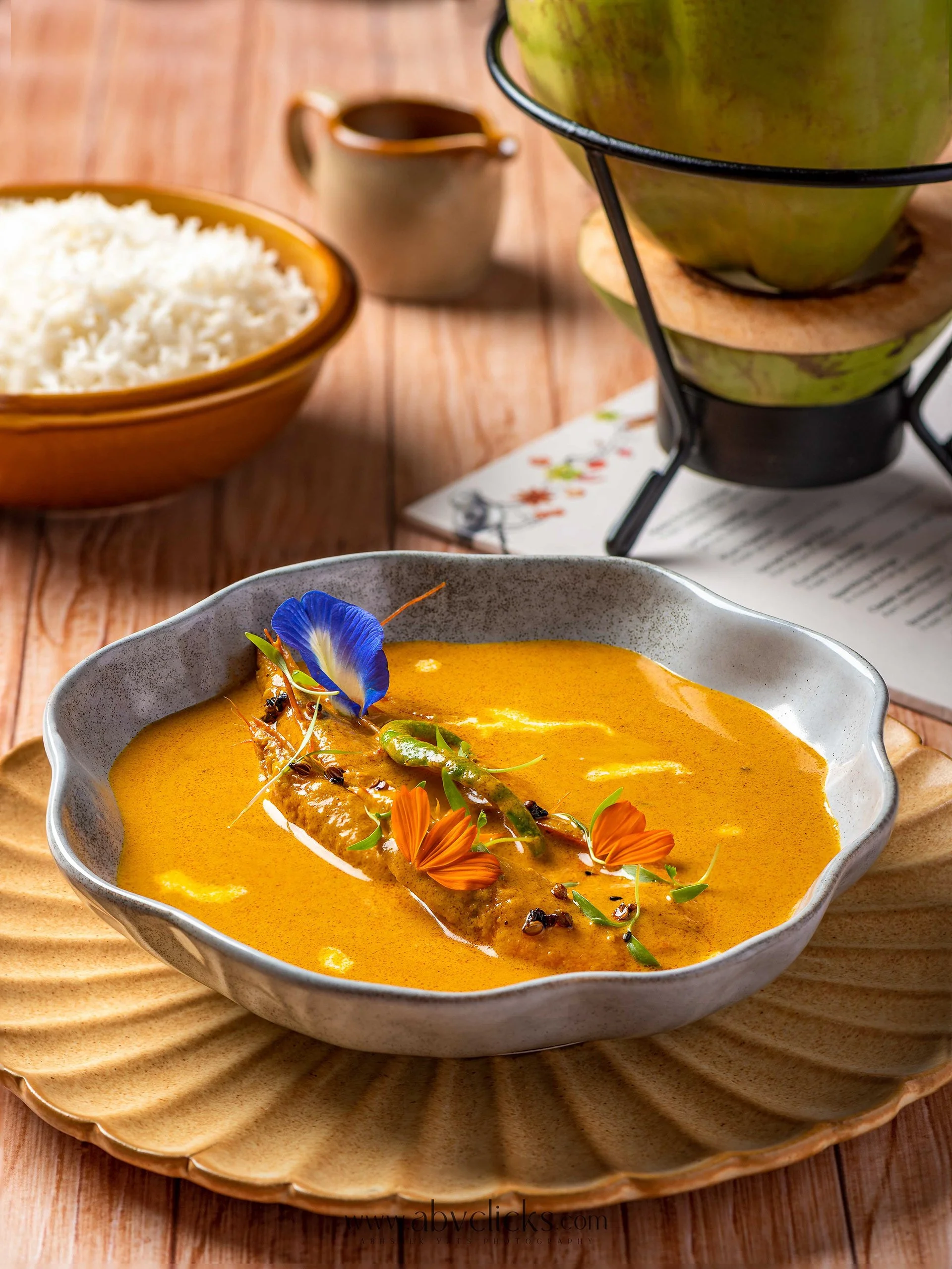 A bowl of yellow curry with a whole fish garnished with edible flowers and herbs, placed on a round wooden plate.