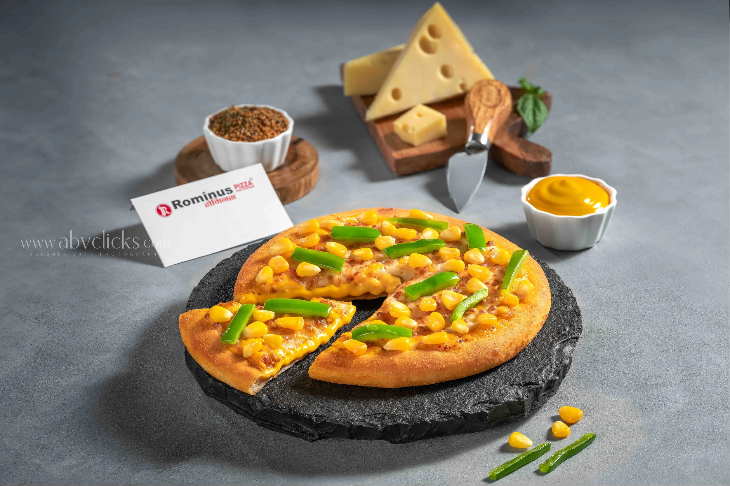 A pizza with cheese, corn, and green peppers on a black slate plate. In the background, there is a small bowl of ground spices, a wedge of Swiss cheese with a cheese knife, and a small bowl of mustard on a wooden cutting board.