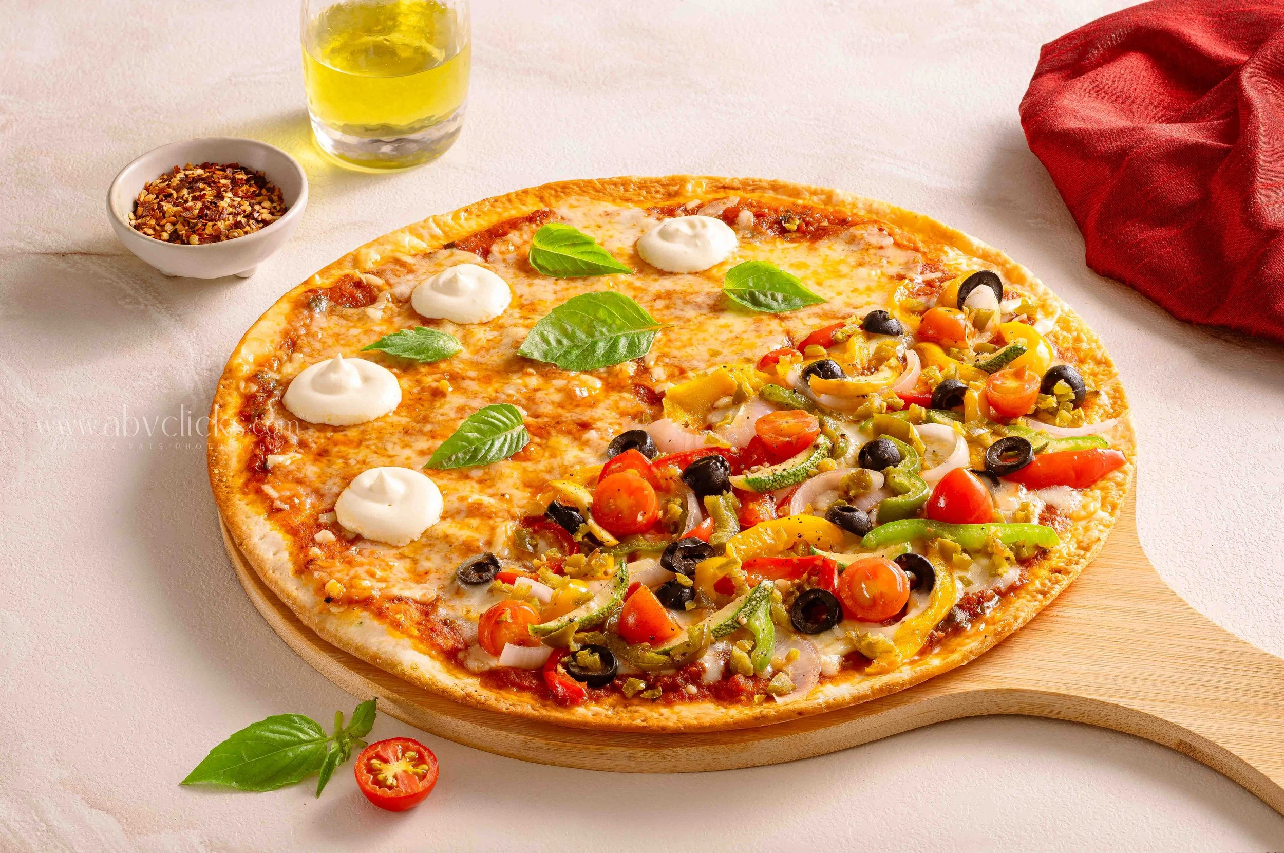 A pepperoni, vegetable, and cheese pizza on a wooden pizza peel, garnished with fresh basil leaves. A glass of yellowish drink and a small bowl of red pepper flakes are in the background.