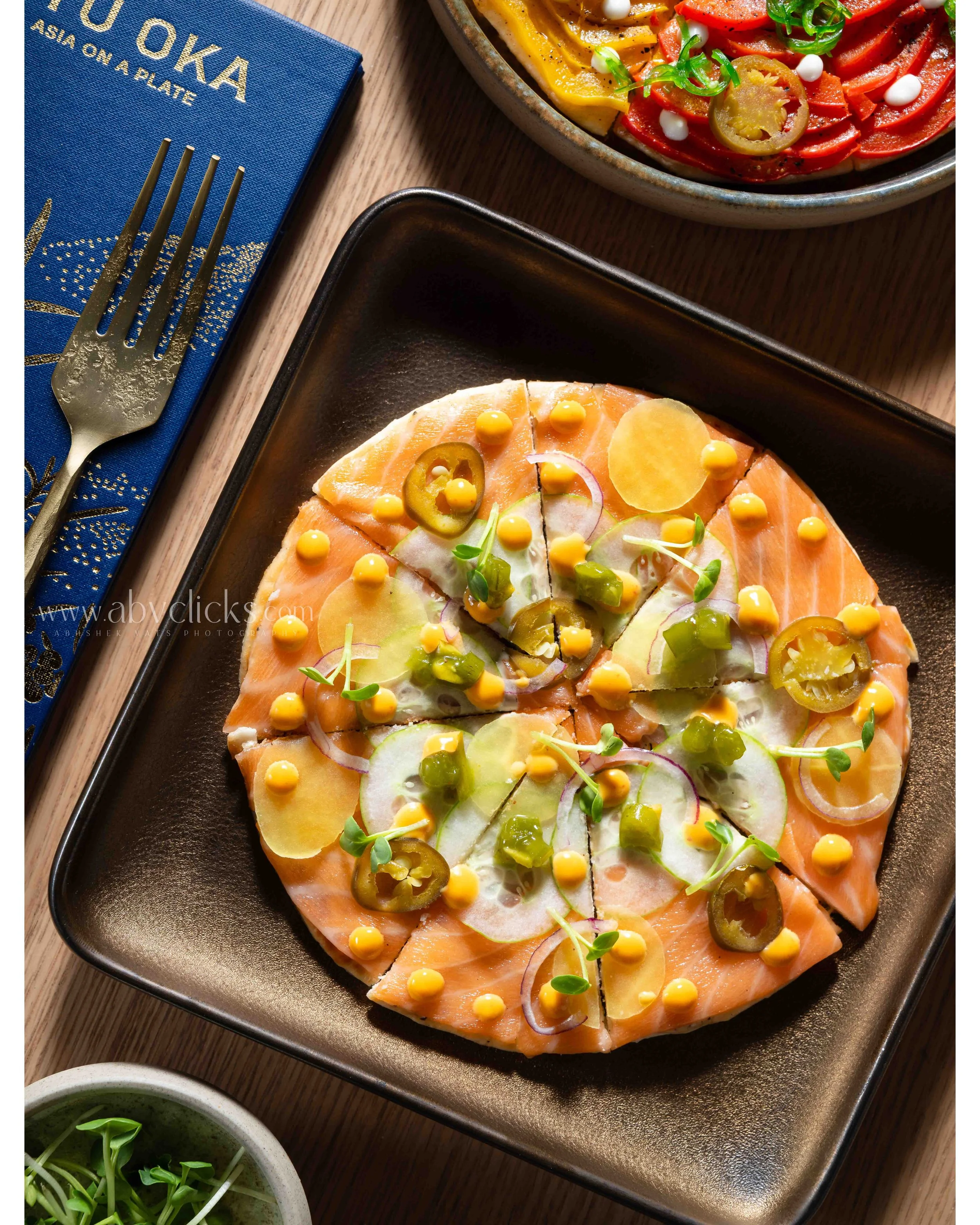 A dinner setting with a plate of smoked salmon appetizer topped with sliced pickles, onions, and microgreens. There is a bowl of colorful sliced peppers and a small bowl of sprouts, along with a menu or placemat that says 'O Tokyo Asia on a Plate' an