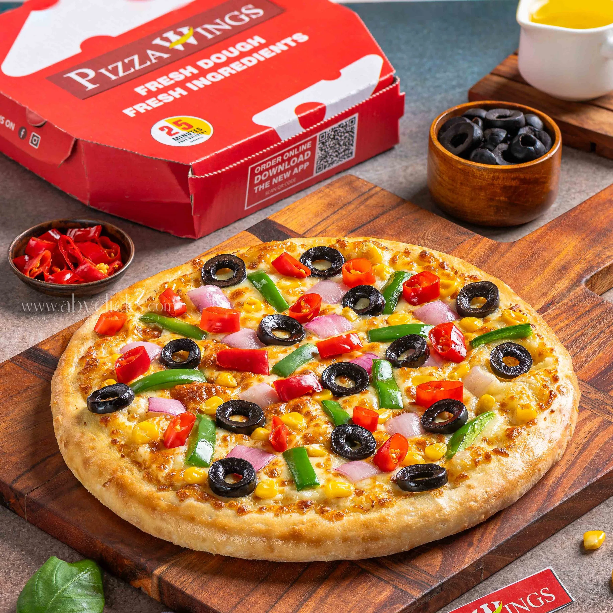 A freshly baked pizza with black olives, red bell peppers, green peppers, onions, corn, and cheese on a wooden board. In the background, there is a pizza box labeled 'Pizza Wings' and some small bowls of toppings and dipping sauces.