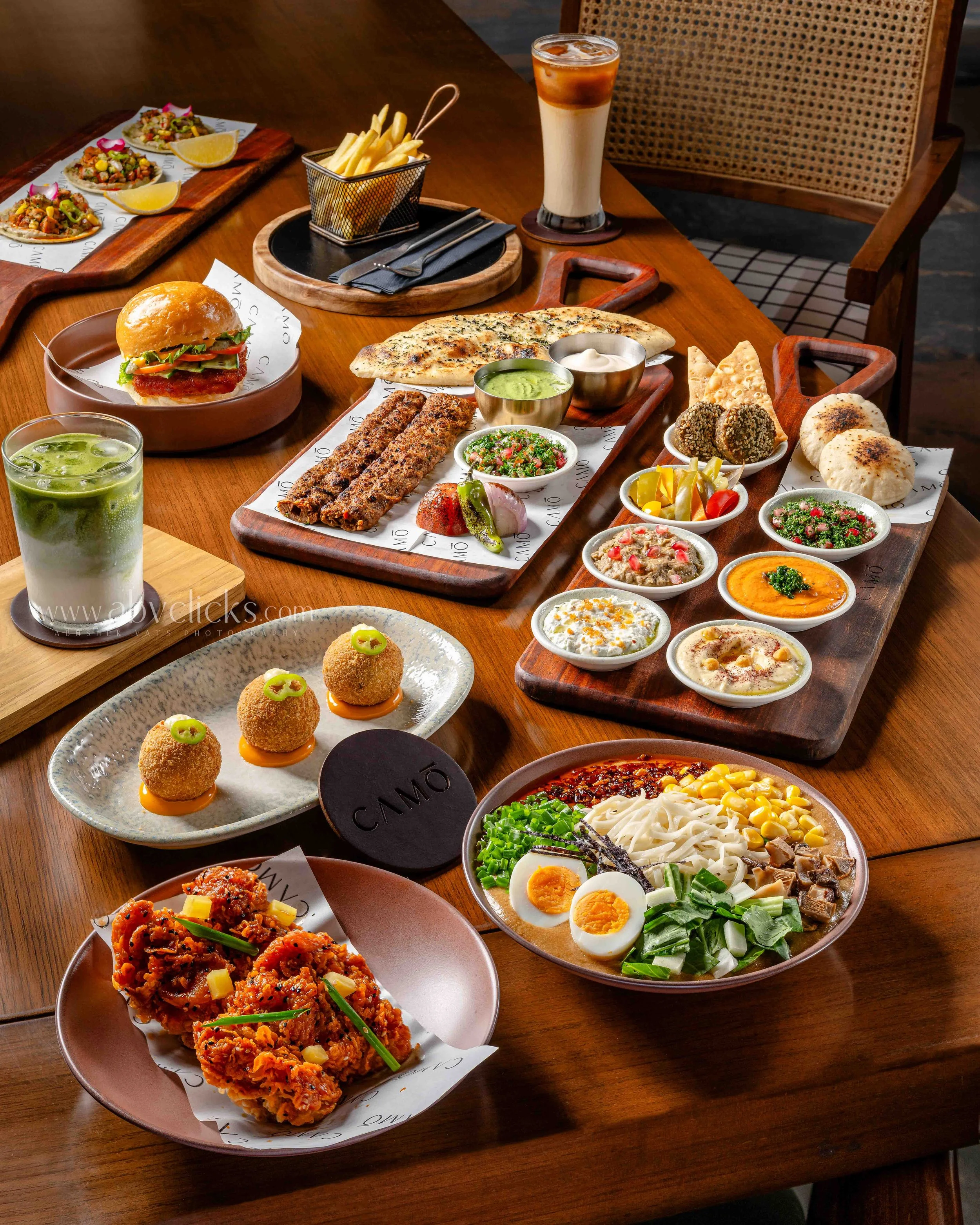 A large wooden table filled with various dishes including fried chicken, salad, grilled meats, naan bread, falafel, dips, and beverages, in a restaurant setting.