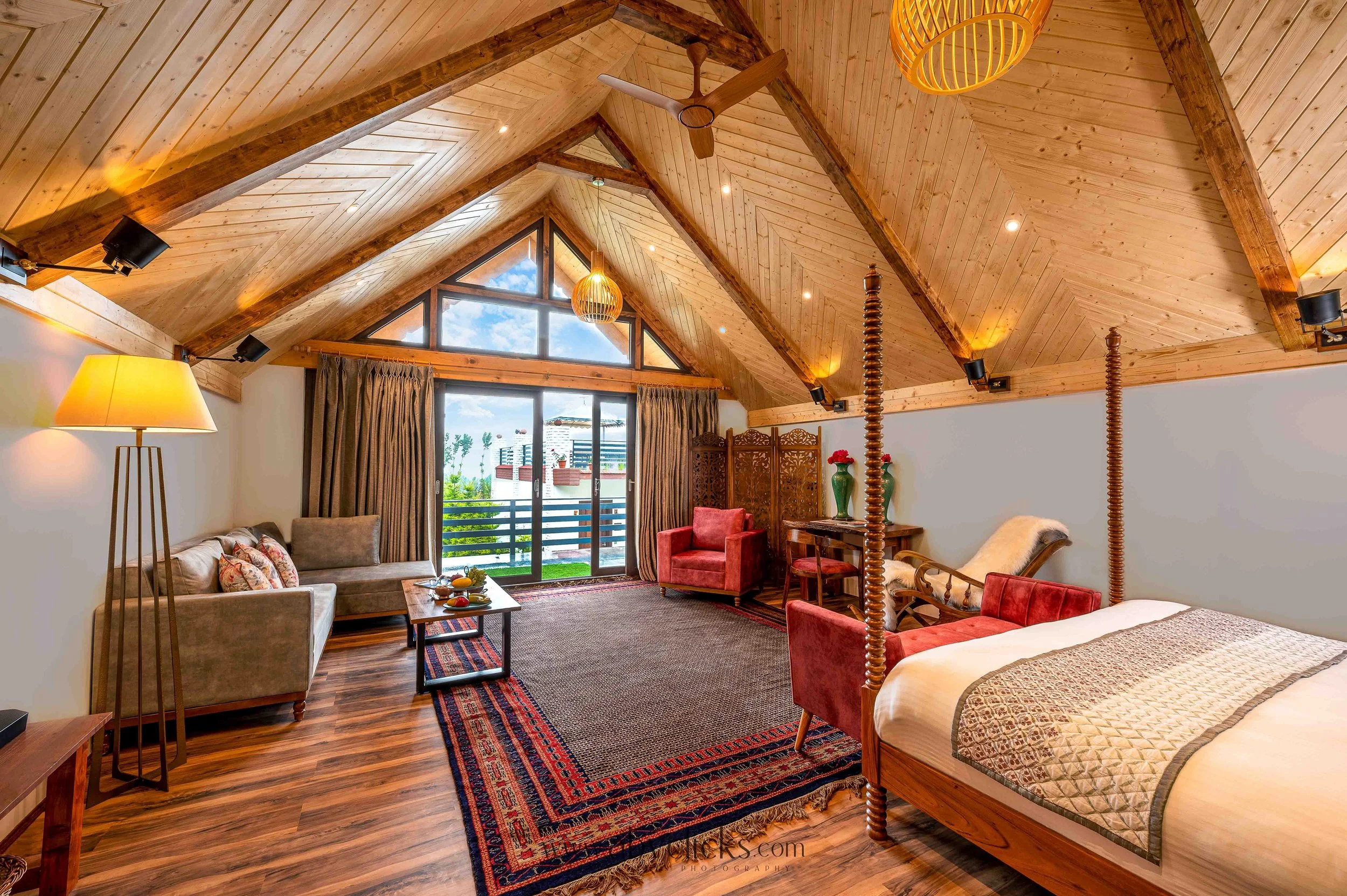 A spacious living room with a high wooden vaulted ceiling, exposed wooden beams, and a ceiling fan. There's a large window and glass doors leading to a balcony with a view of trees and rooftops. The room has a mix of seating including a beige sofa, r