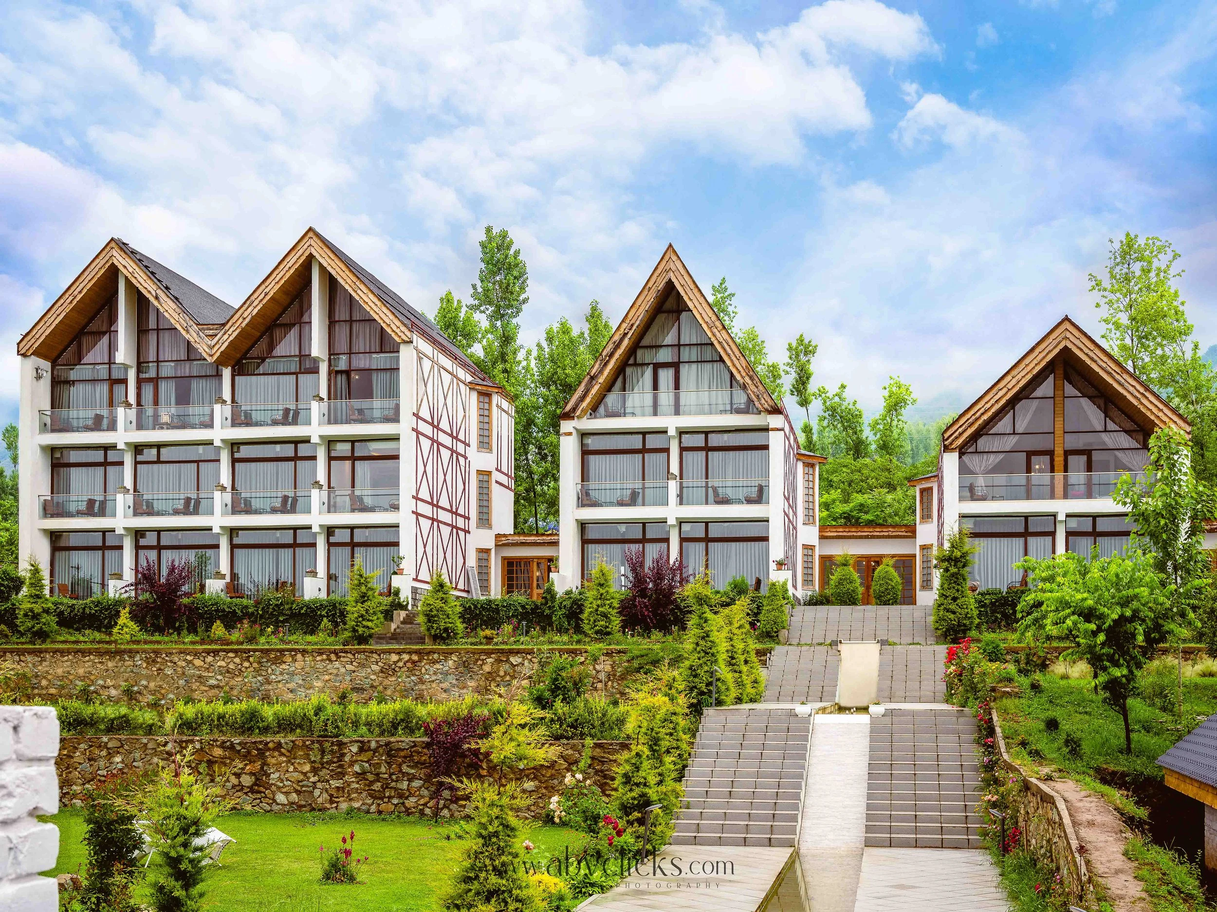 A residential property with three modern A-frame cabins with glass balconies surrounded by lush greenery, trees, and a stone and concrete staircase leading to the entrance, under a partly cloudy sky.