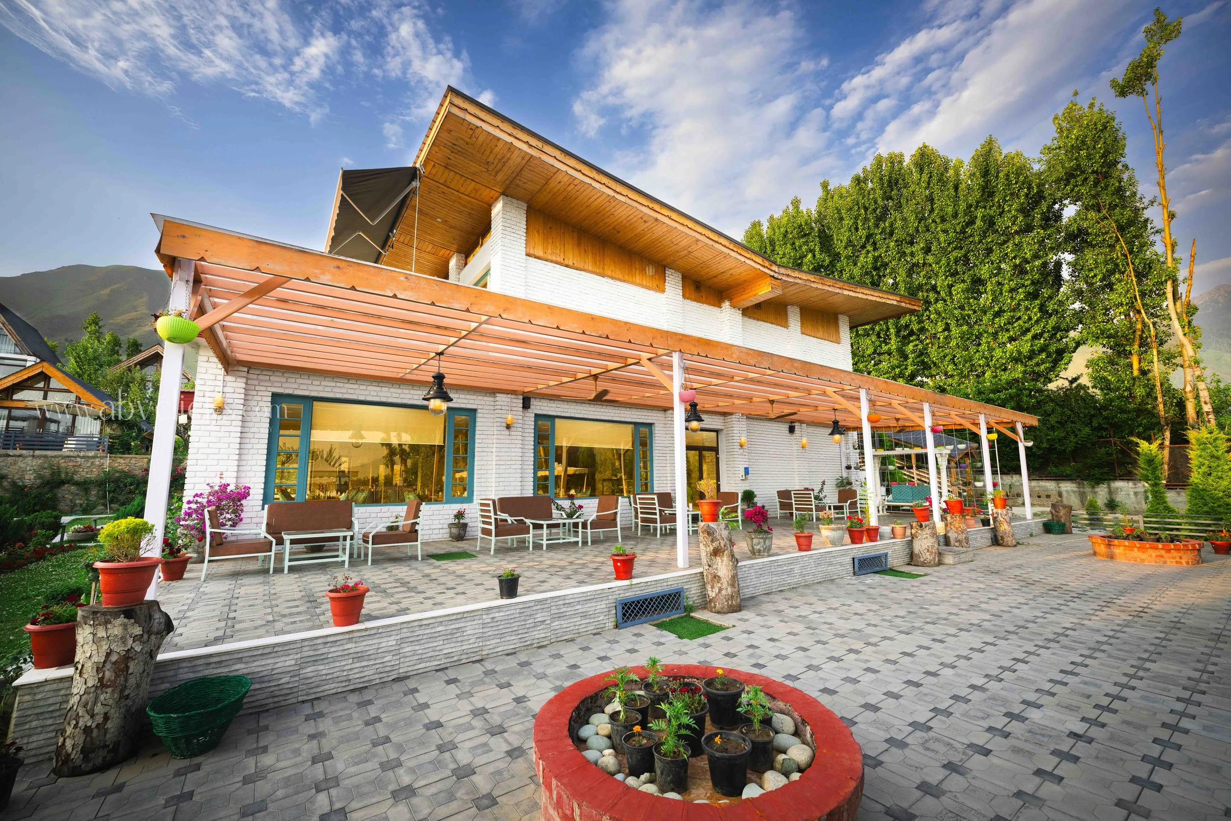 A modern two-story house with white brick walls and a sloped wooden roof. The house features large glass windows and a spacious outdoor patio with patio furniture, potted plants, colorful lanterns, and decorative elements. The surrounding yard has a 