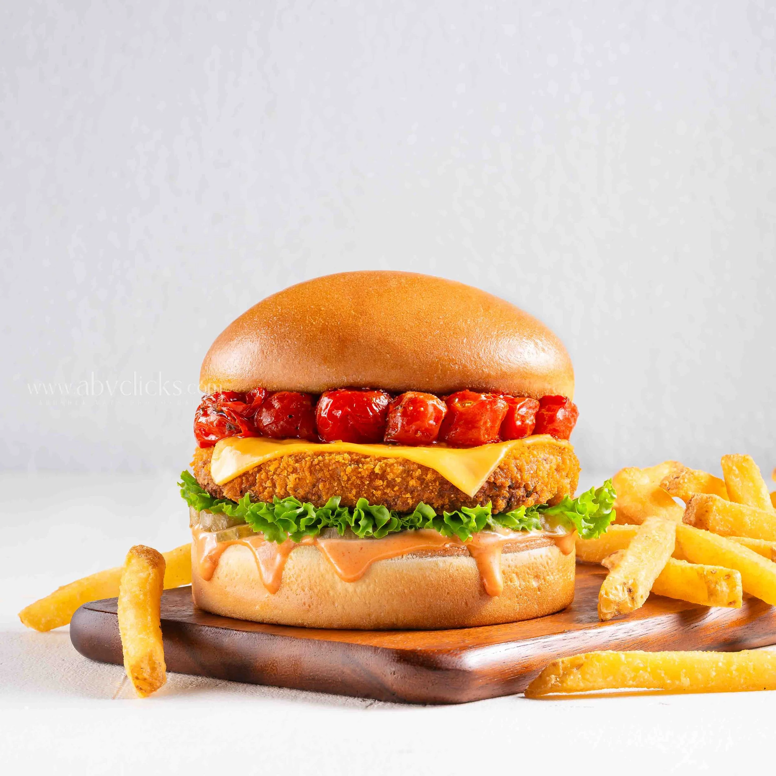 A close-up of a sandwich with fried chicken fillet, lettuce, cheese, and sauce in a bun, on a wooden board, with fried potato chips on the side.