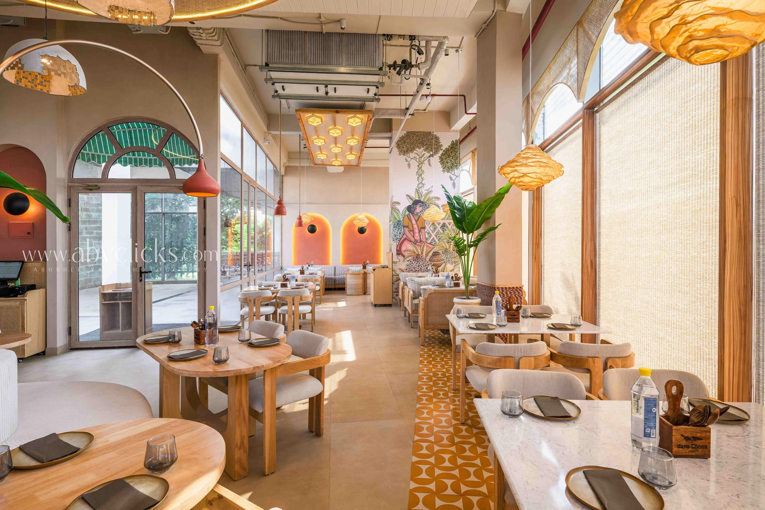 Interior of a modern restaurant with wooden furniture, large windows with woven blinds, hanging light fixtures, and a colorful mural on the wall.