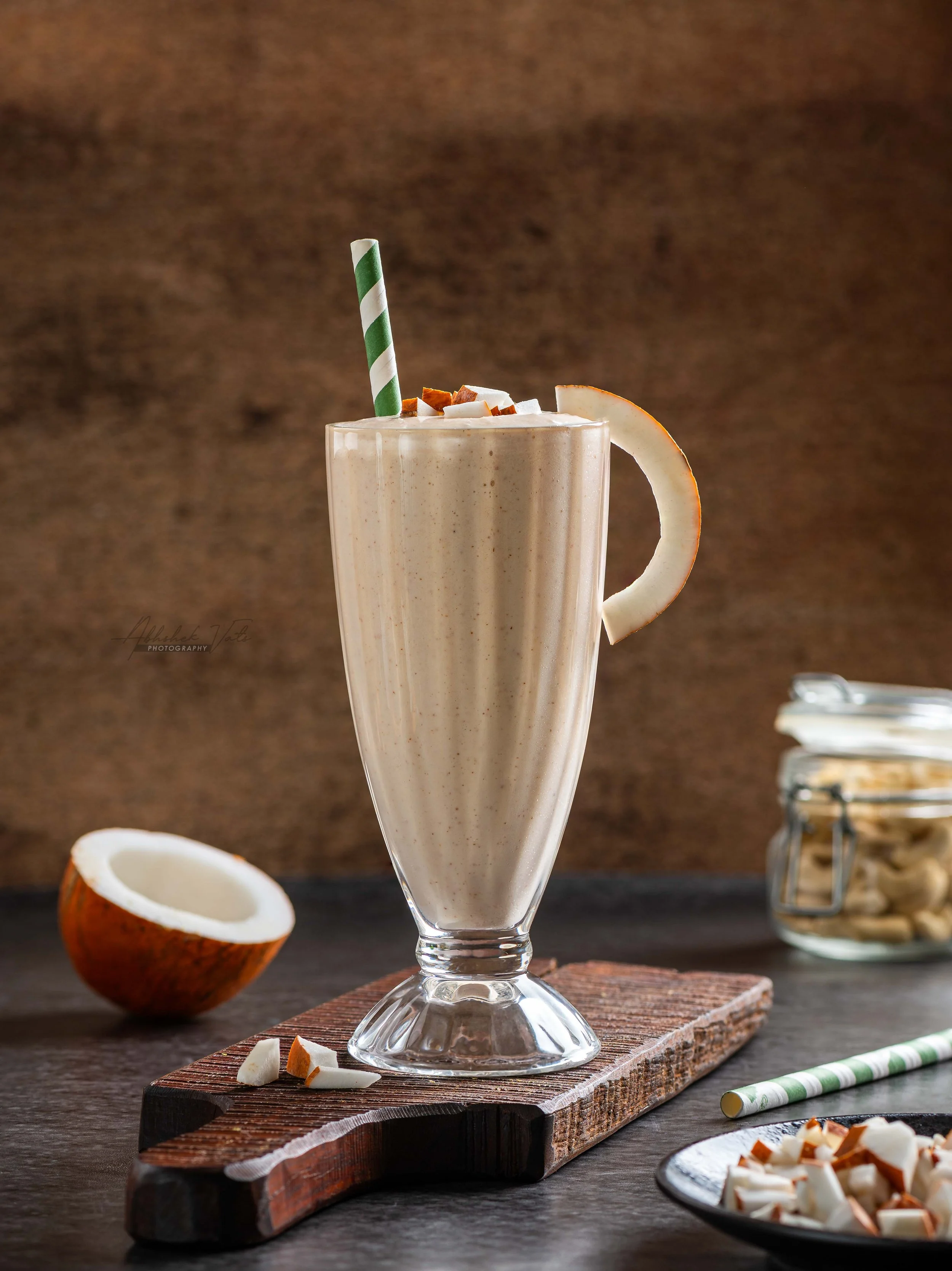 A tall glass of almond milkshake topped with whipped cream, coconut flakes, and chopped almonds, with a green and white striped straw. The glass is on a wooden board on a dark table, with a halved coconut and a plate of chopped coconut and almonds ne