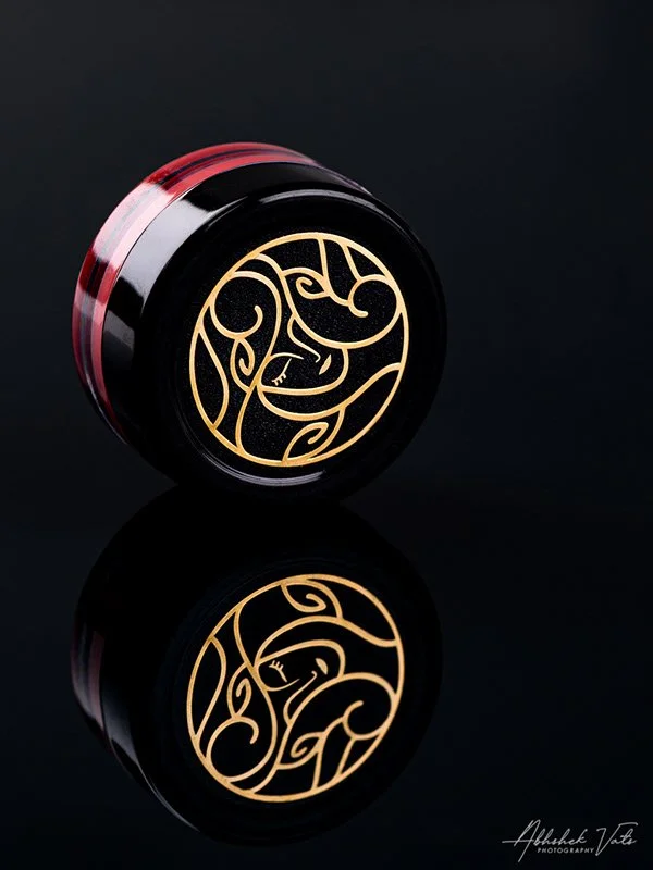 A black jar with a gold tribal design on its lid, placed on a reflective surface against a black background.