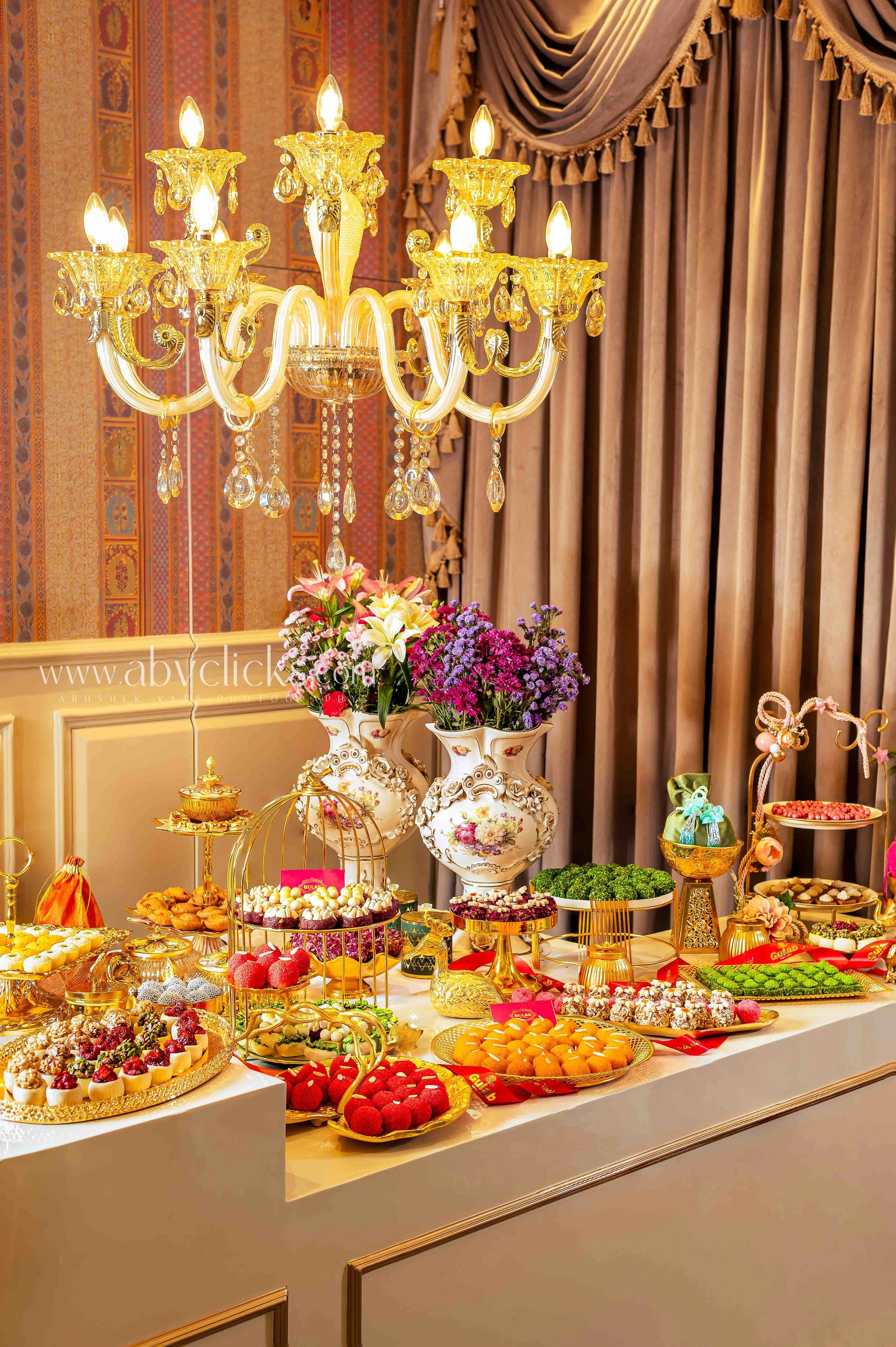 A luxurious dessert table decorated with a large, ornate chandelier, pink and purple flowers in vases, and an array of colorful desserts and sweets on golden trays and plates, set against a background of elegant curtains and patterned wallpaper.