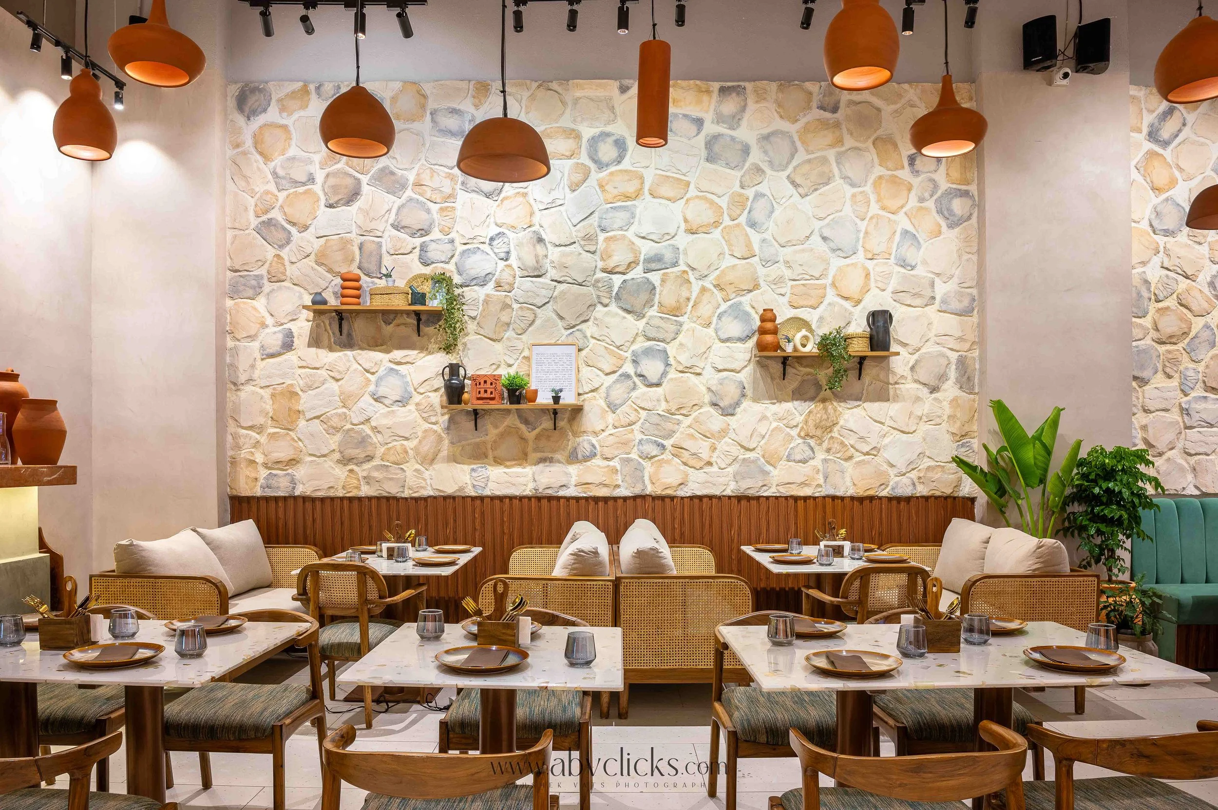 Interior of a modern restaurant with a stone accent wall, wooden shelves with decorative items, and tables set with plates and glasses, featuring hanging pendant lights.
