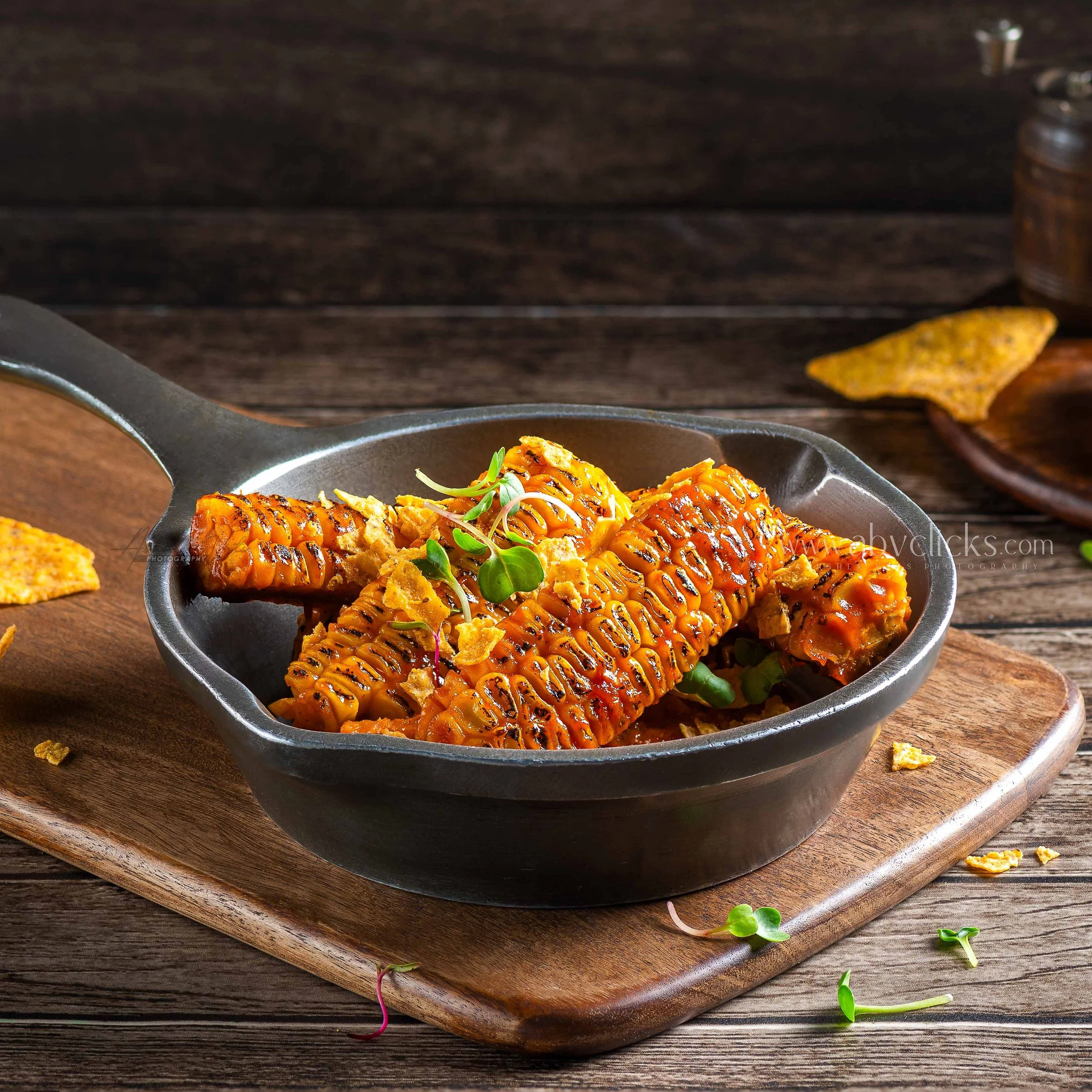 Grilled salmon fillet with grill marks, topped with orange sauce and microgreens, served on a black skillet. A yellow tortilla chip is on the side, on a wooden cutting board, with scattered microgreens and a small jar in the background.