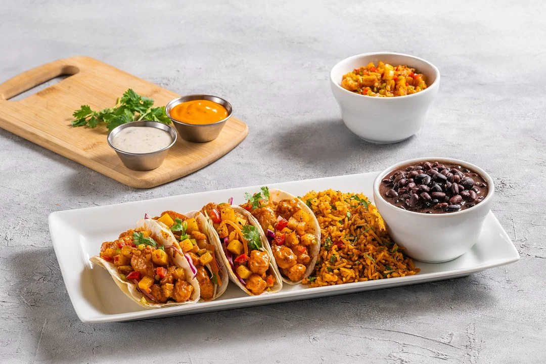 Plate with three tacos filled with chicken, vegetables, and cilantro, along with rice and black beans, topped with small bowls of sauce and a small bowl of mixed vegetables.